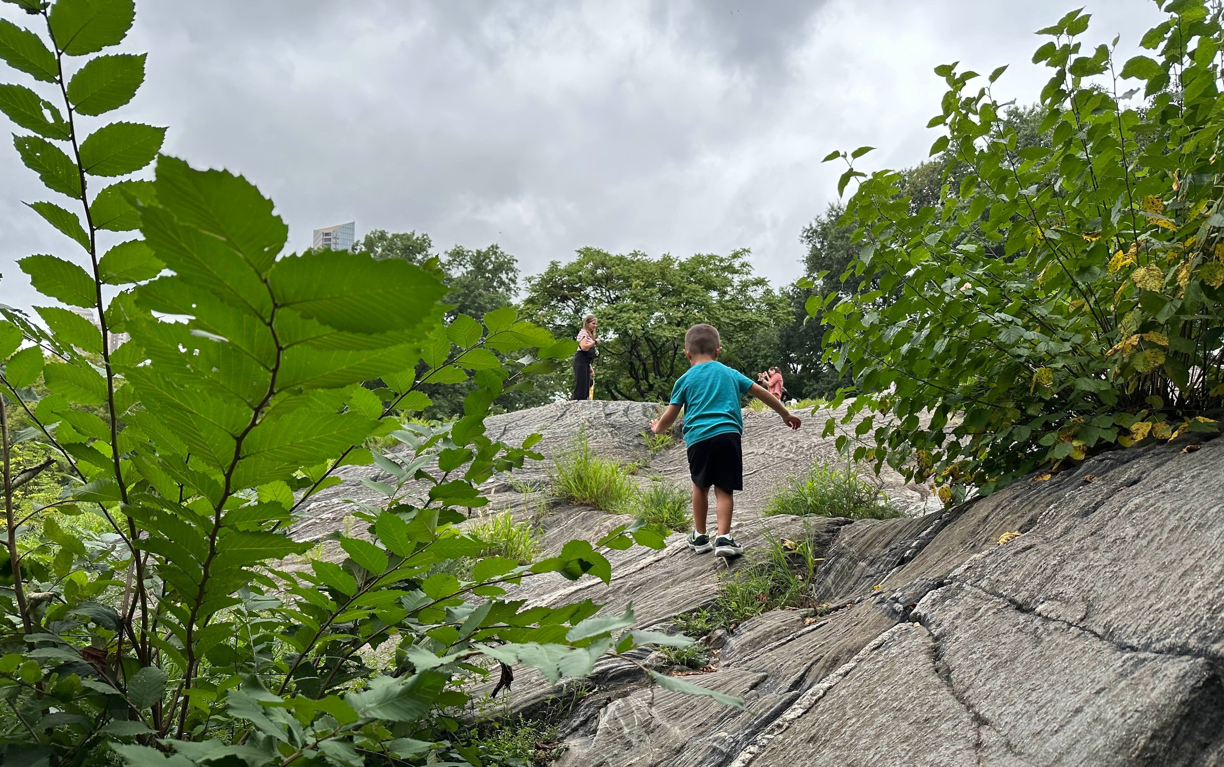 A child runs up a rocky incline towards two adults in a natural setting under overcast skies.