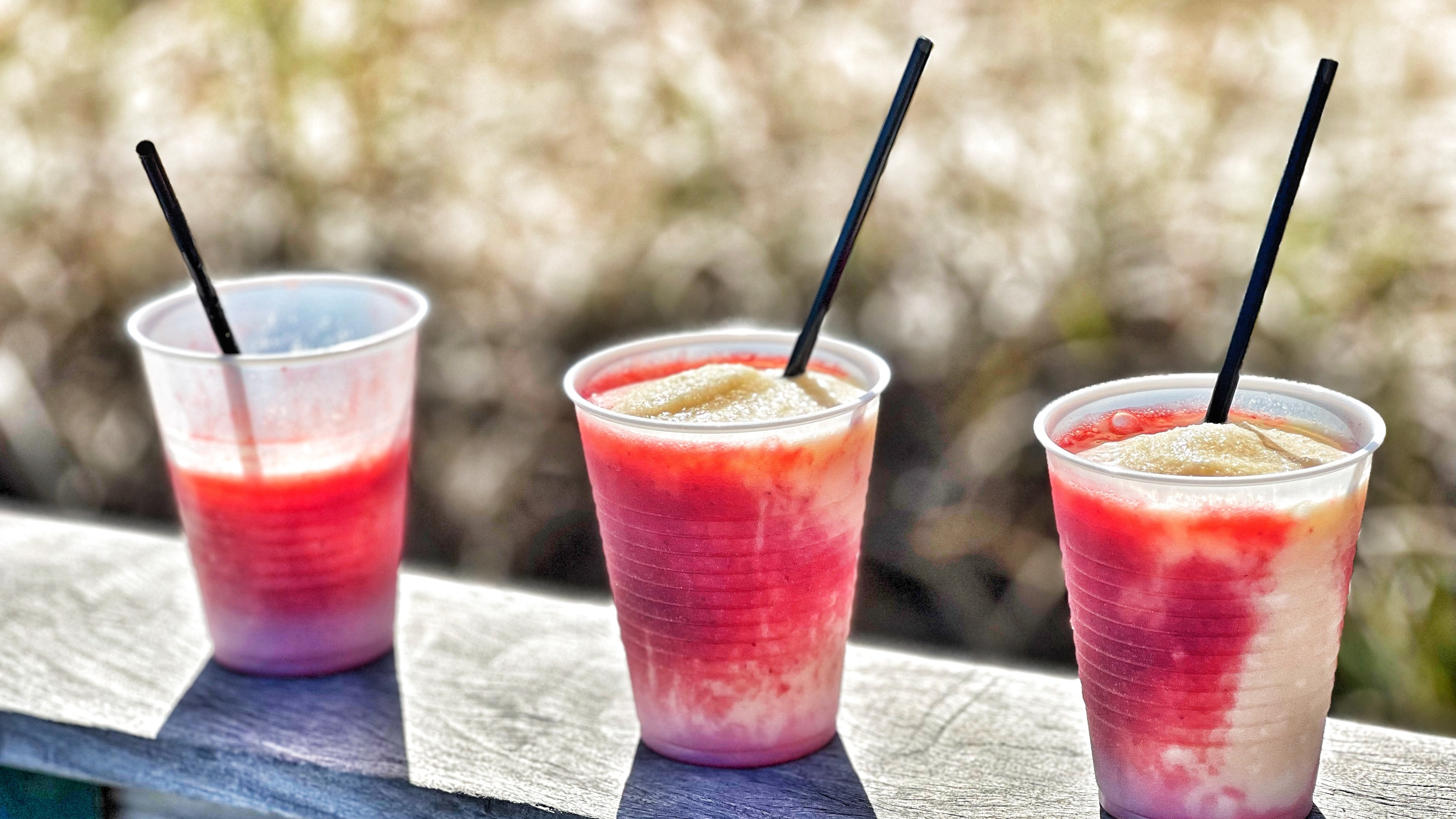 Three red frozen drinks with straws balanced on a wooden deck railing.
