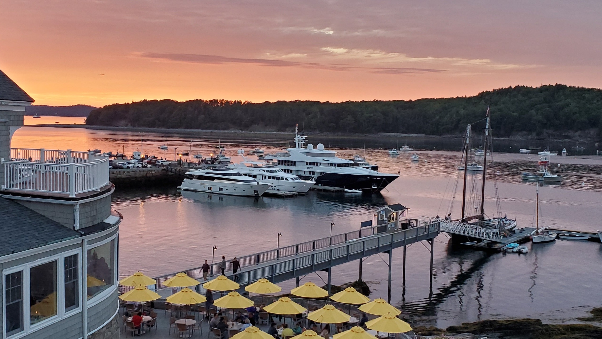 The image shows a scenic sunset view over a harbor with boats, featuring a building with a deck and yellow umbrellas.