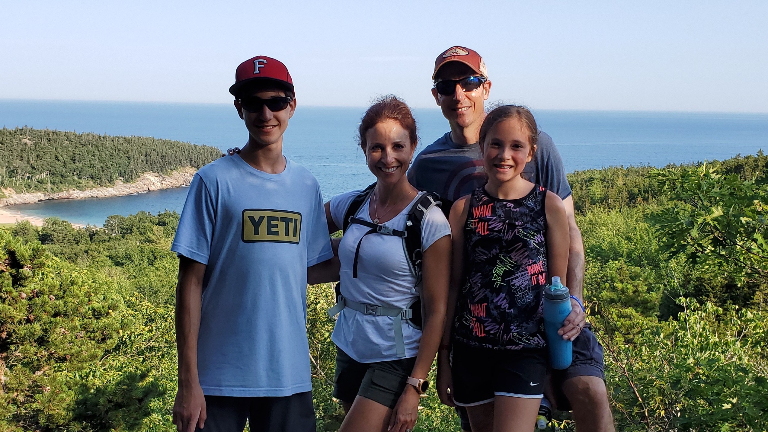 The image shows four individuals standing in an elevated area with a view of a forested region and a beach in the background.