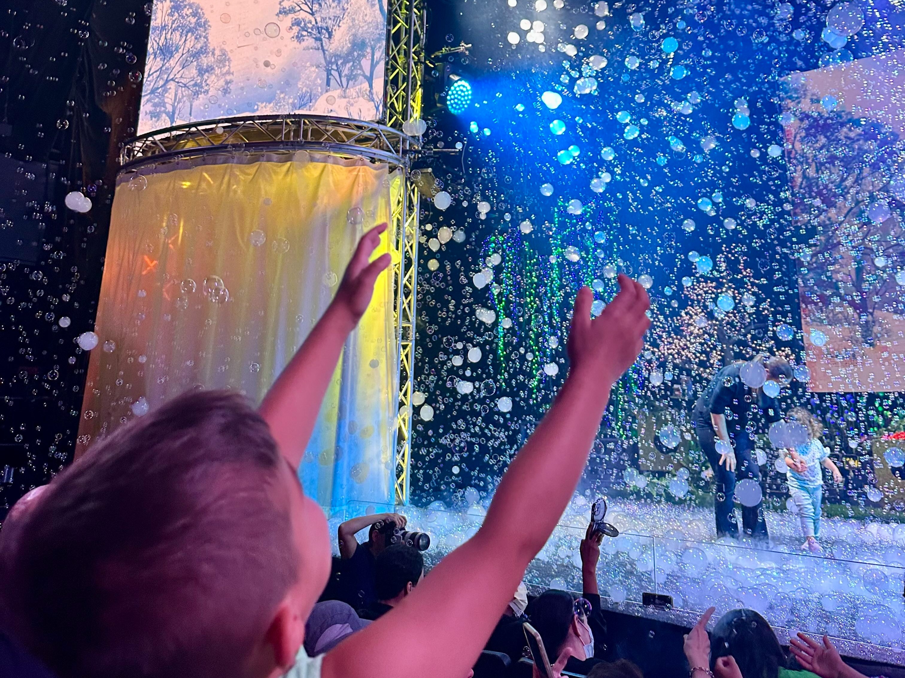 A child reaches out towards a stage filled with floating soap bubbles during an event.