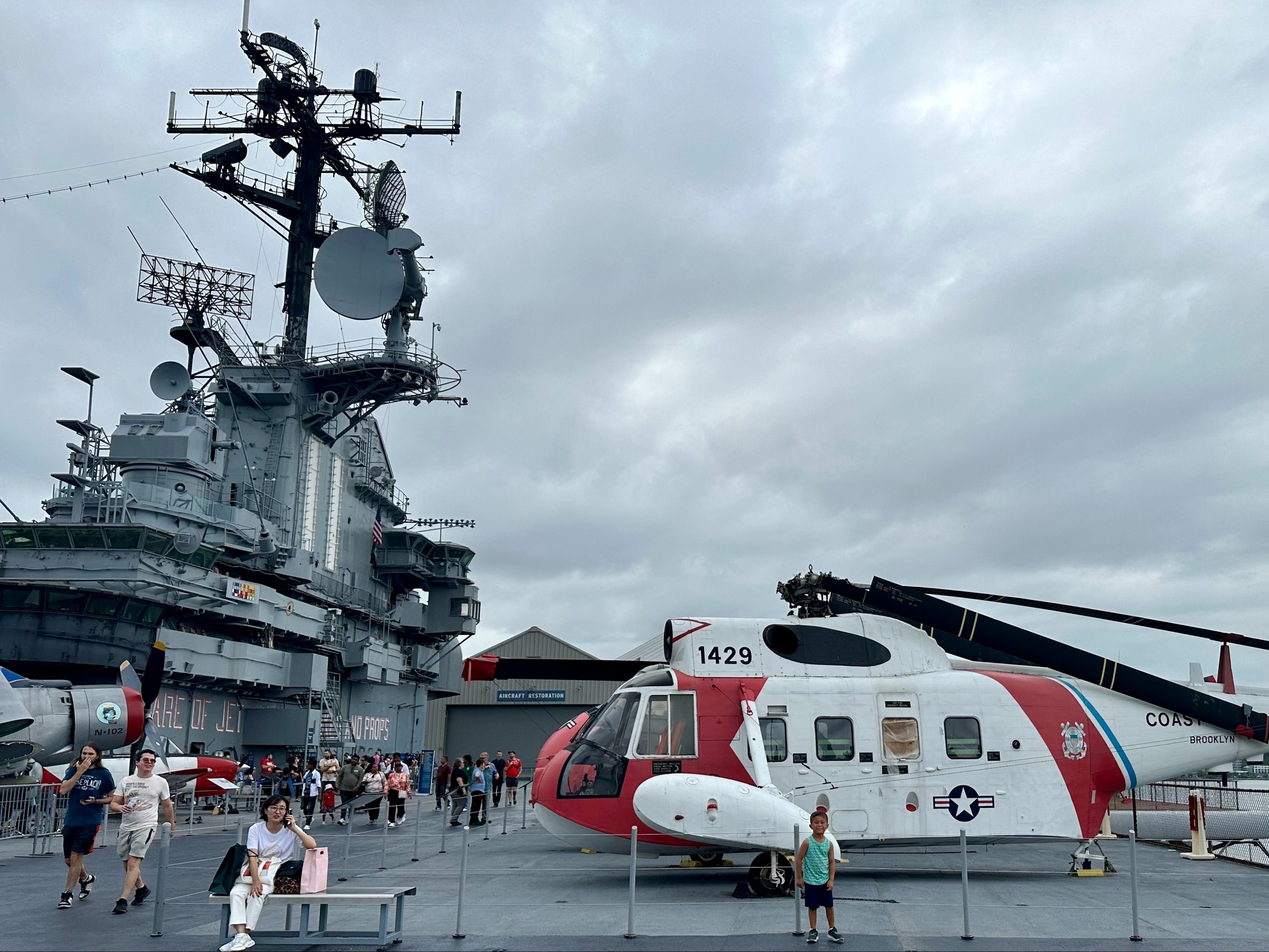 A helicopter is parked on the deck of an aircraft carrier, with people walking around under a cloudy sky.