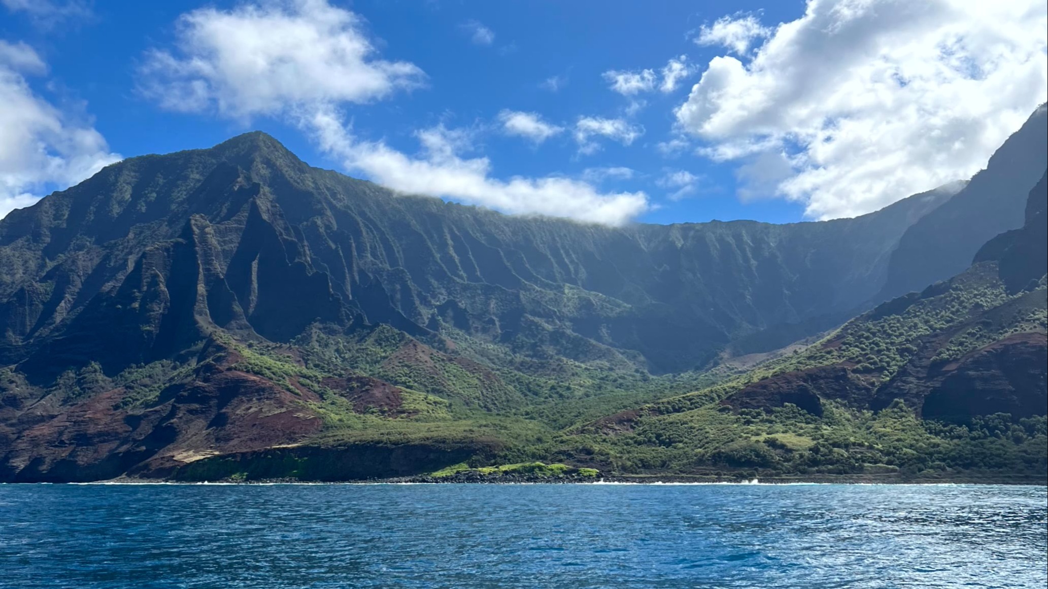 A coastal landscape with a body of water in the foreground and a steep, green cliff in the background under a blue sky with clouds.