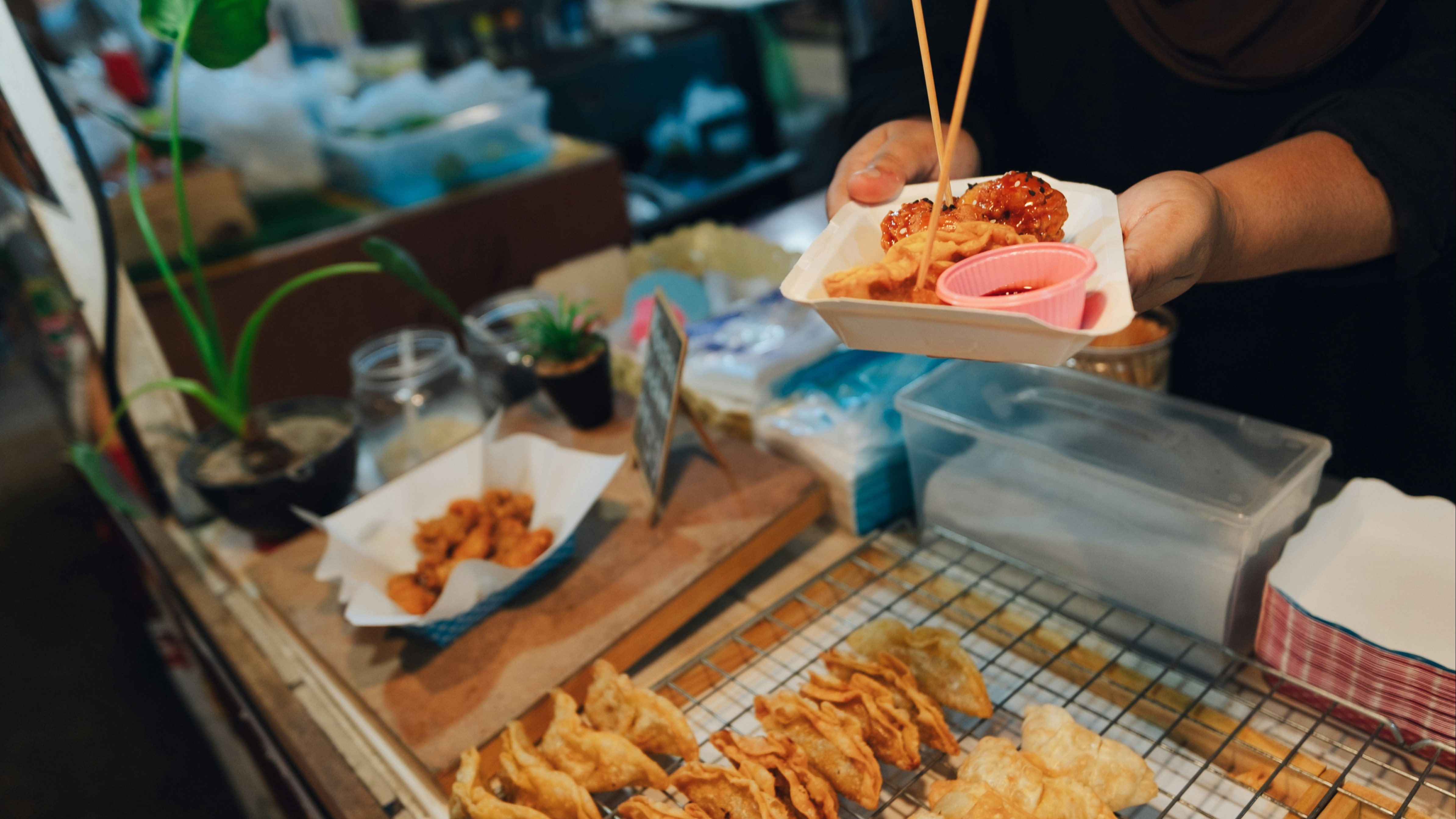 A person holds a plate of food with chopsticks at a market stall, showcasing various fried foods.