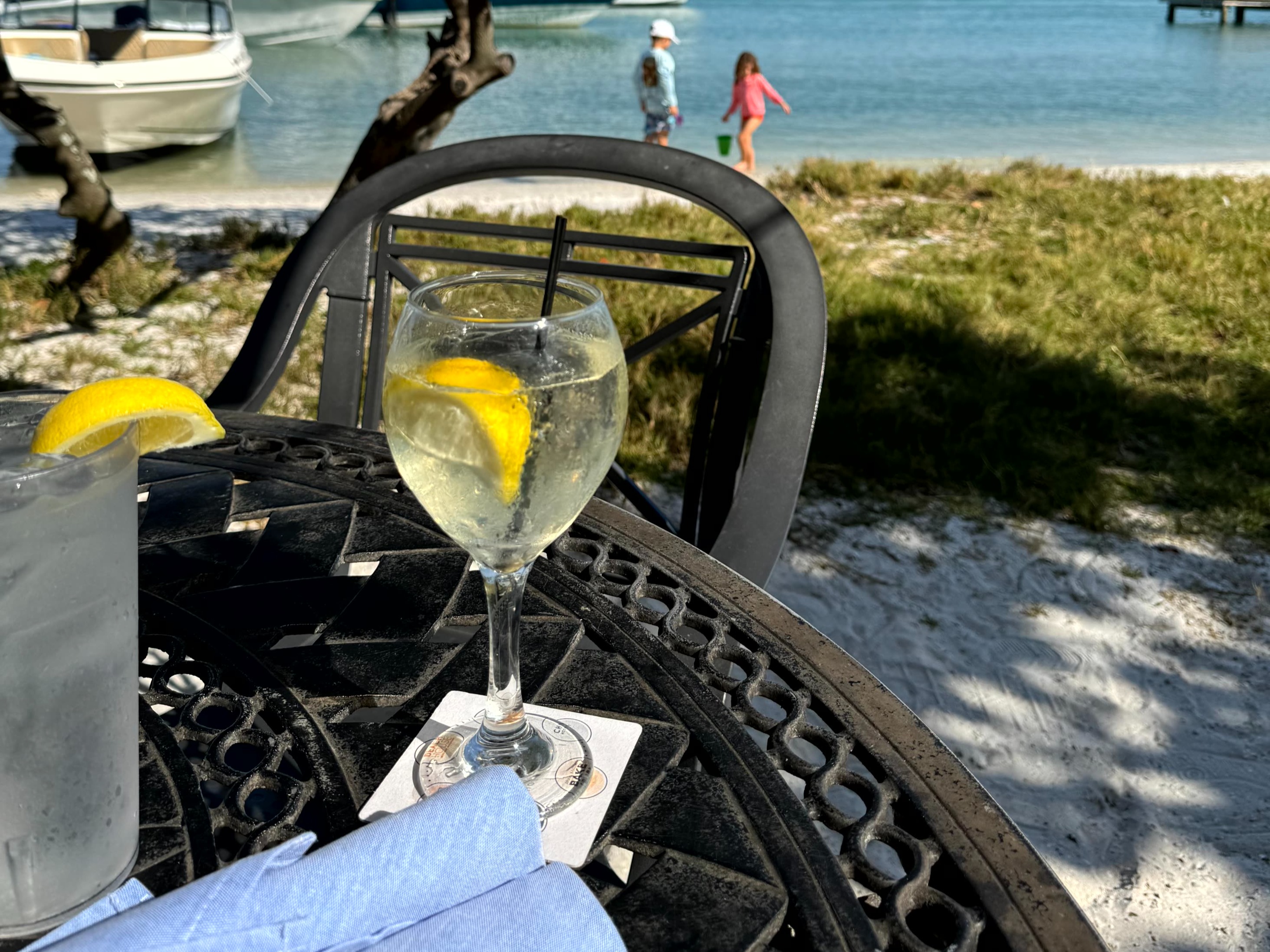 A refreshing drink with a lemon slice sits on an outdoor table by the water, with a boat and person blurred in the background.