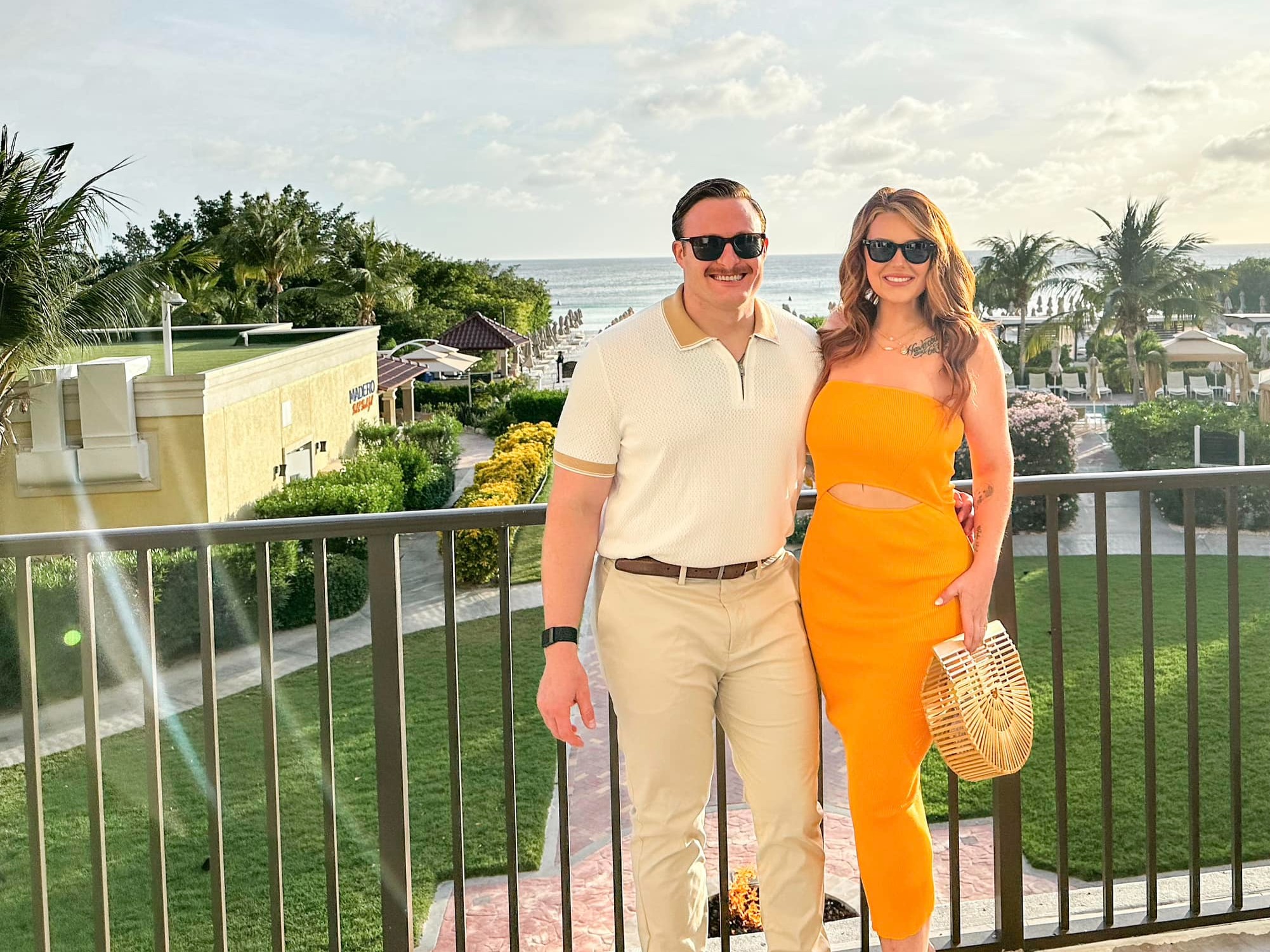 The image shows two individuals wearing sunglasses and standing on a balcony with a beachside view.