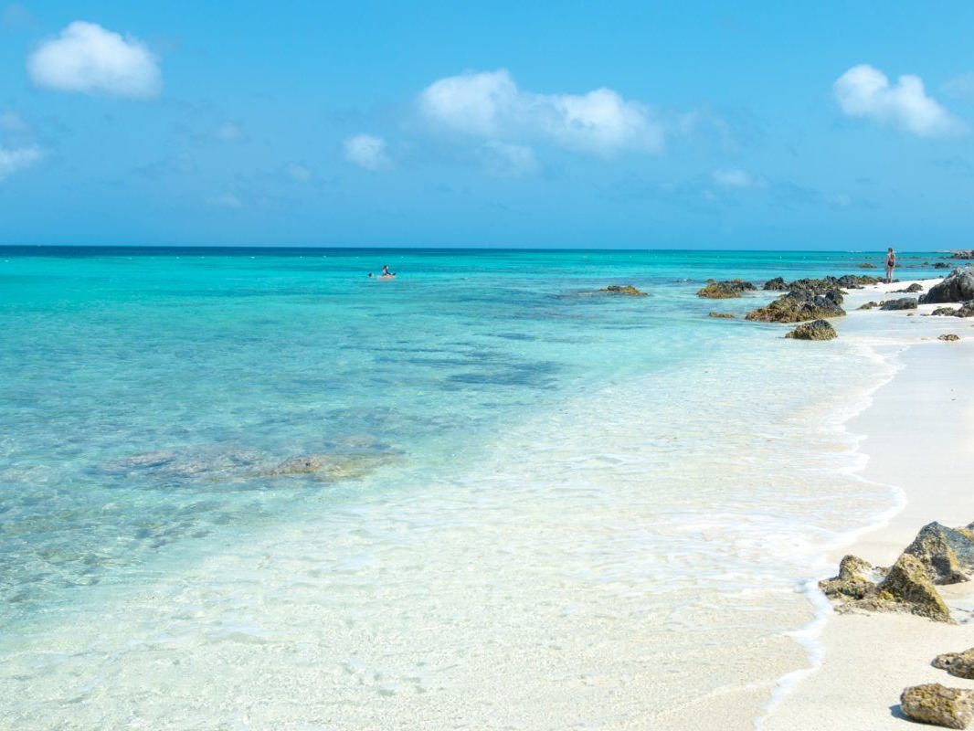 The image shows a serene beach with clear turquoise waters, white sand and scattered rocks under a blue sky with fluffy clouds.