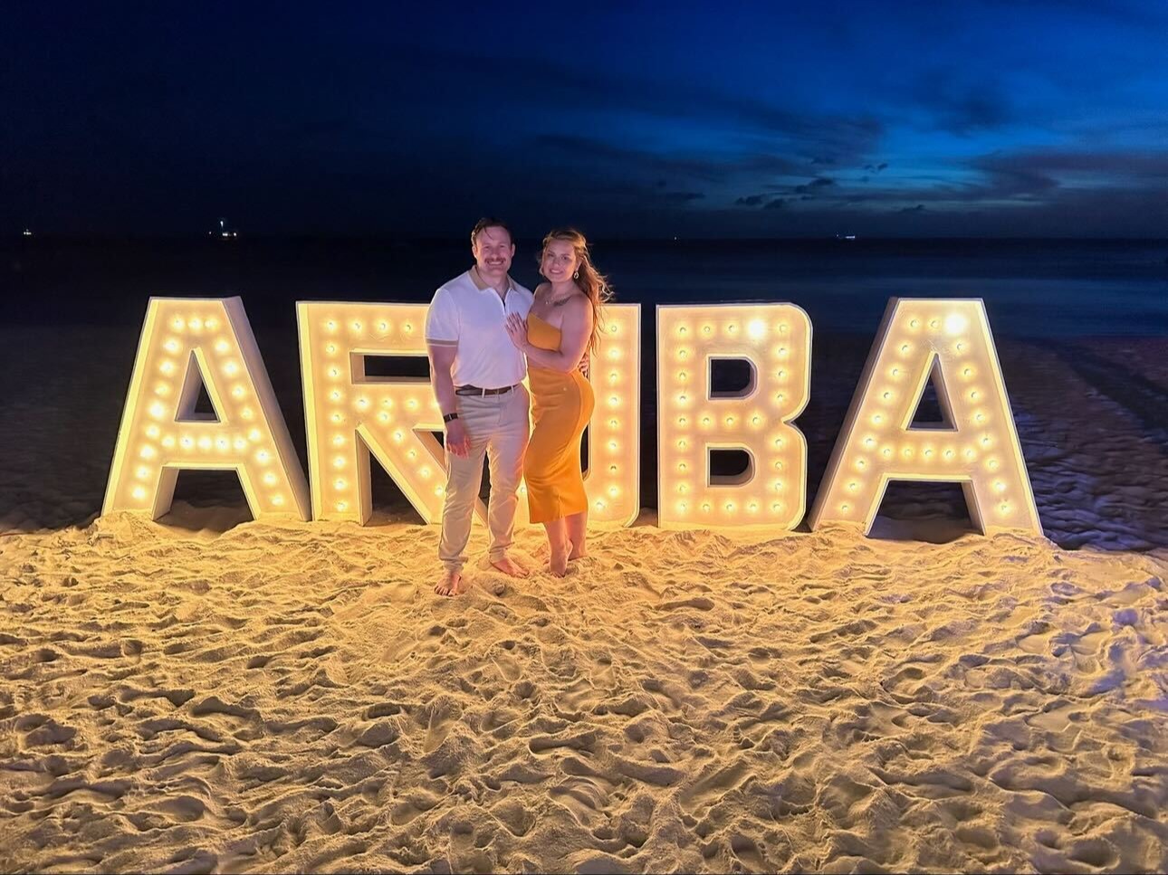 The image shows two people standing on a beach at twilight in front of large illuminated letters spelling “ARUBA”.