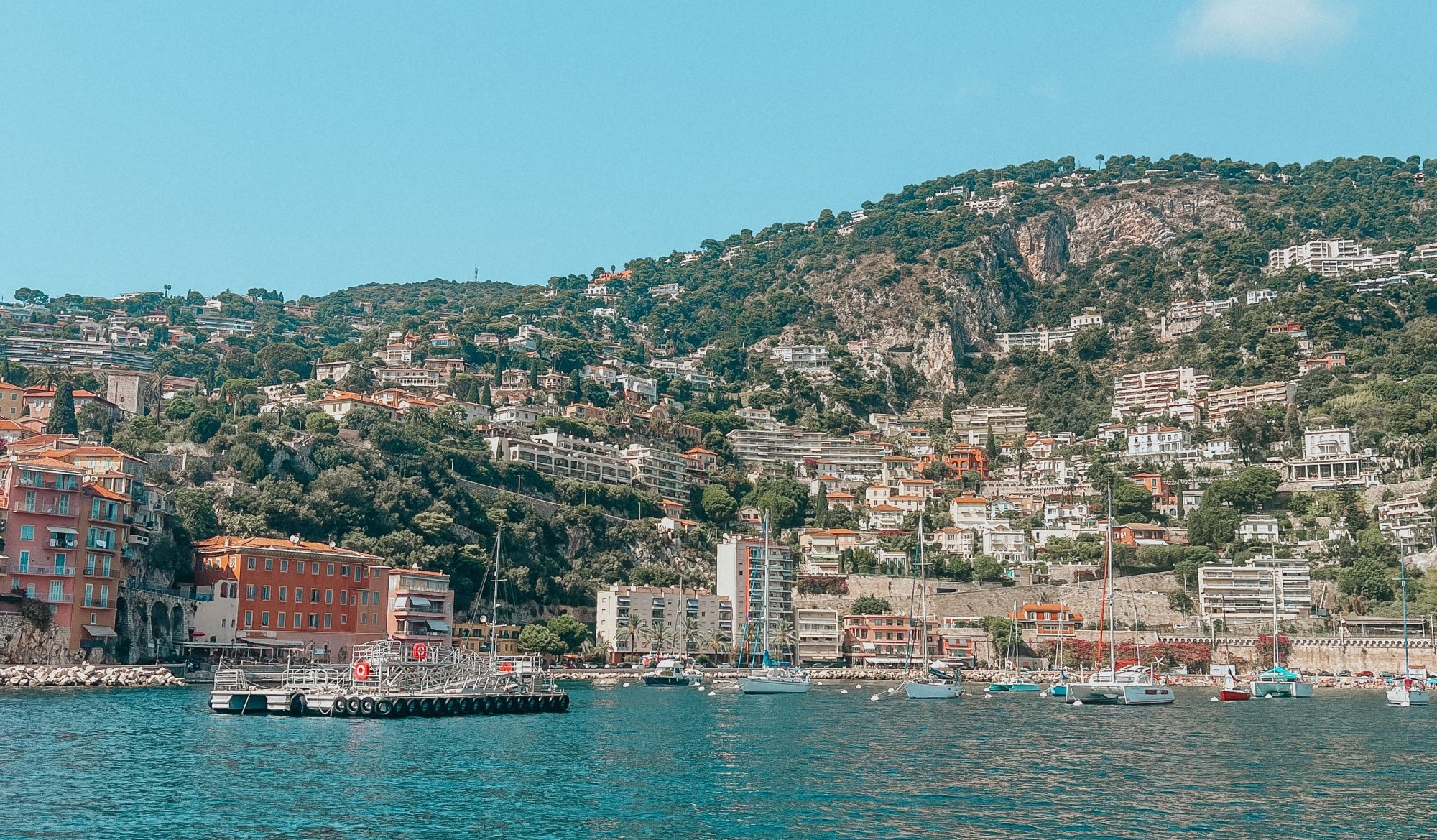 View of the lake with a boat floating towards the coastline and buildings on a tree covered mountainside with a blue sky above.