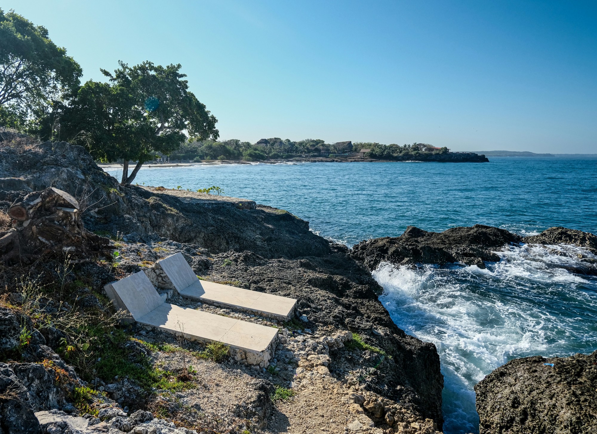 Two lounge chairs on a rocky beach with waves crashing below them.