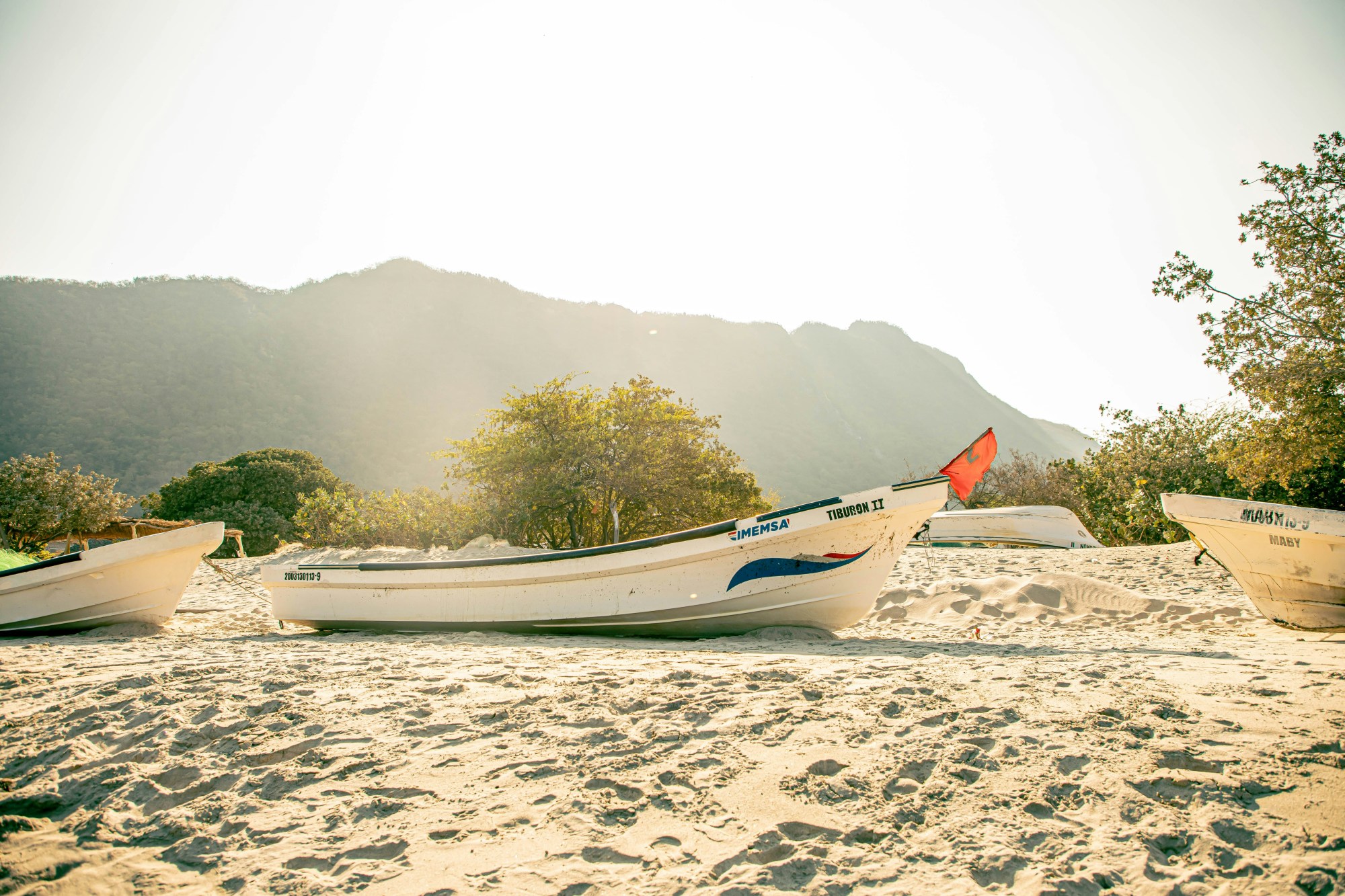 A view of white boats lined up on a sandy beach with a large mountain behind them and the sun shining above.