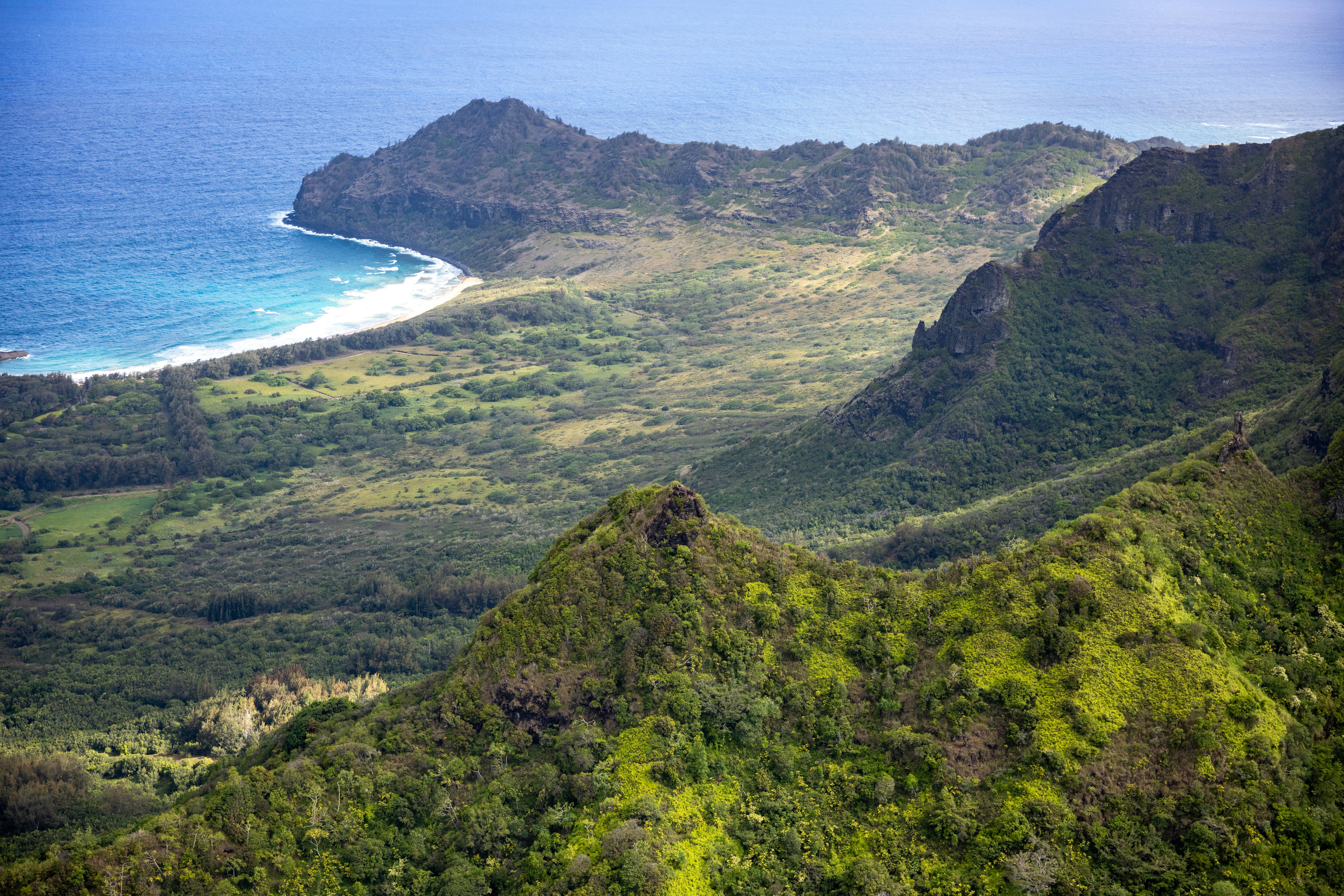 An aerial view of the coastline and green mountains on a hazy sunny day.