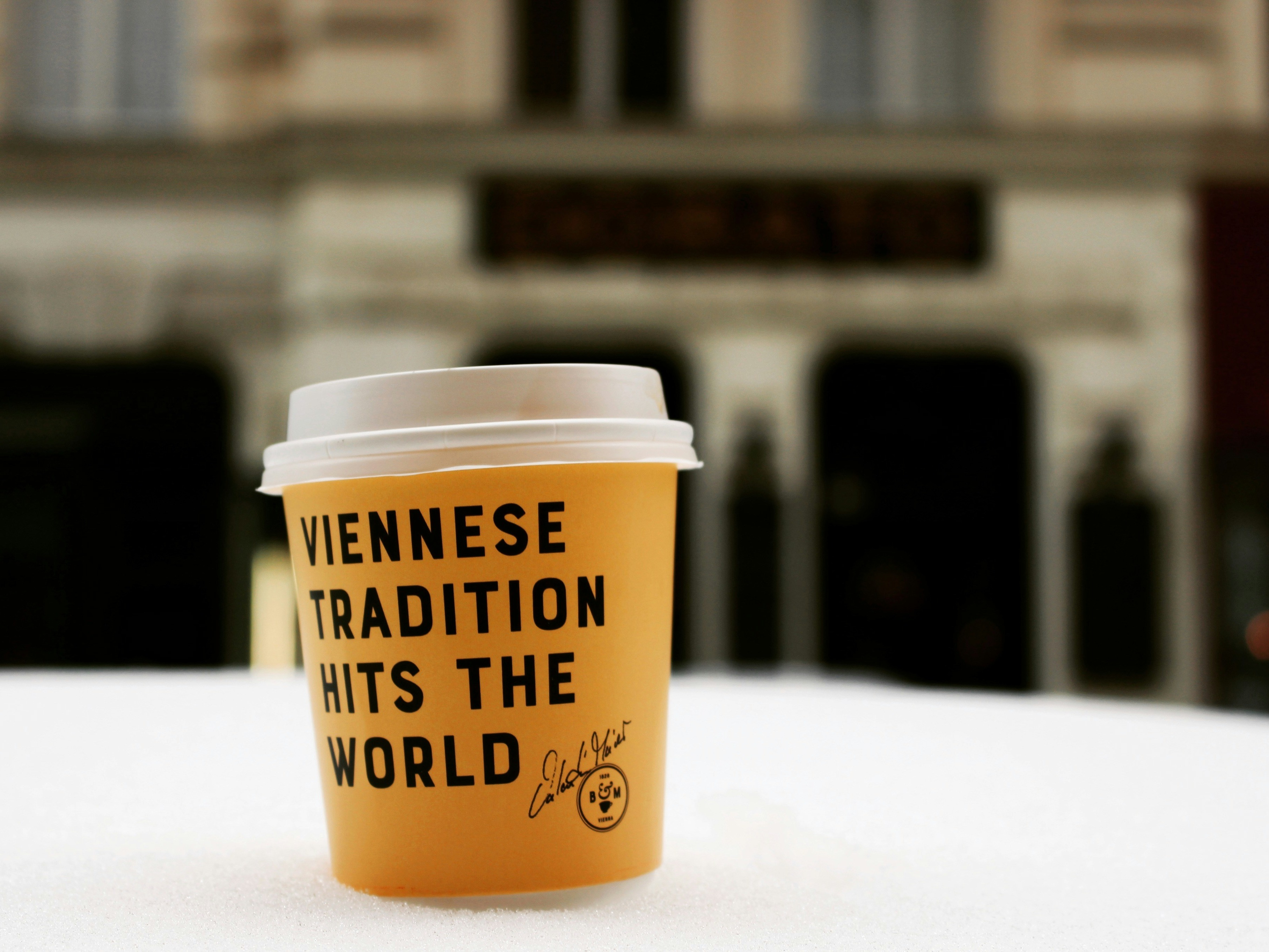 A coffee cup with the text “VIENNESE TRADITION HITS THE WORLD” against a backdrop of blurred buildings.