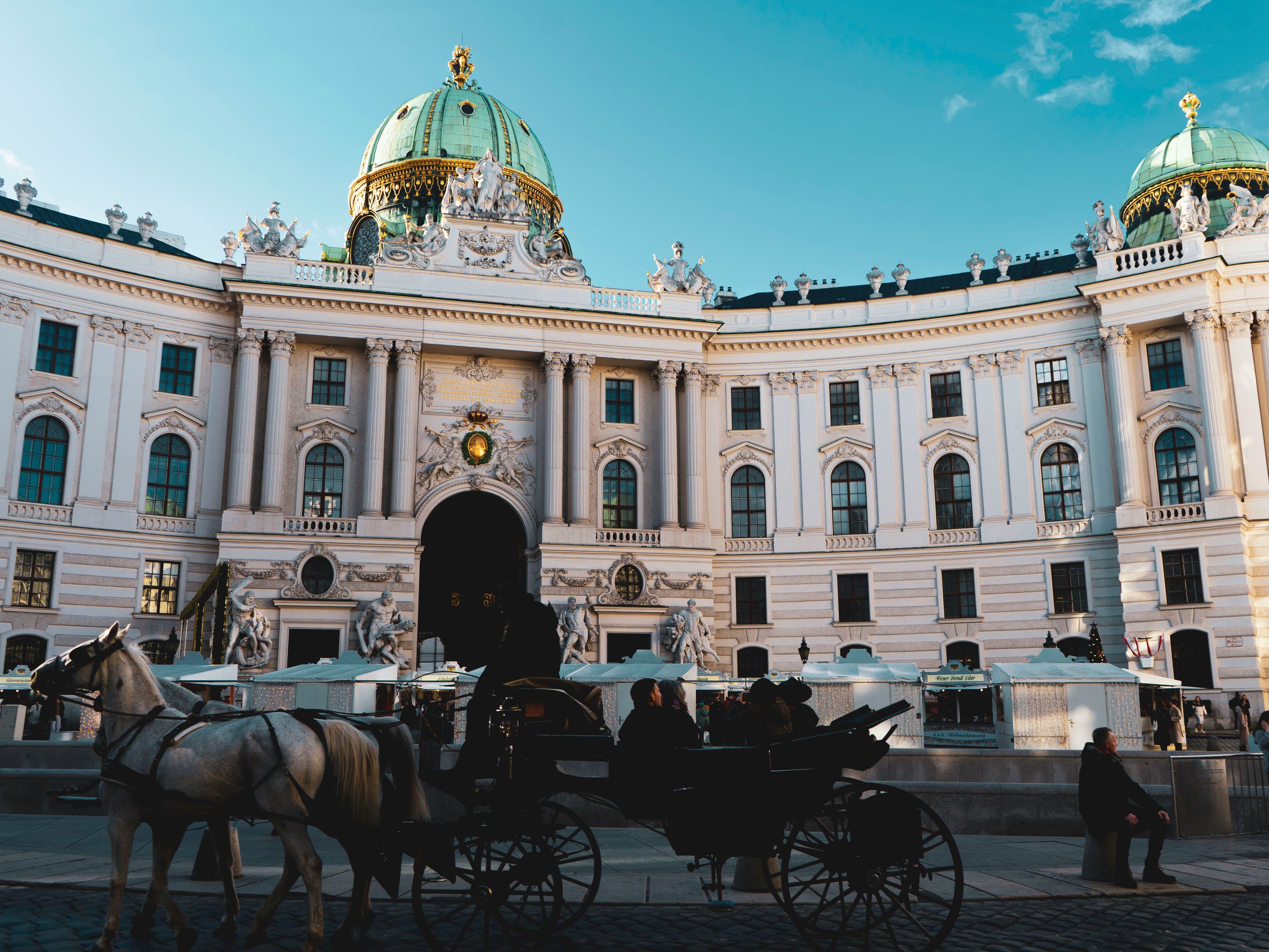 A grand, ornate building with a dome and sculptures, a horse-drawn carriage in the foreground, and people milling about.