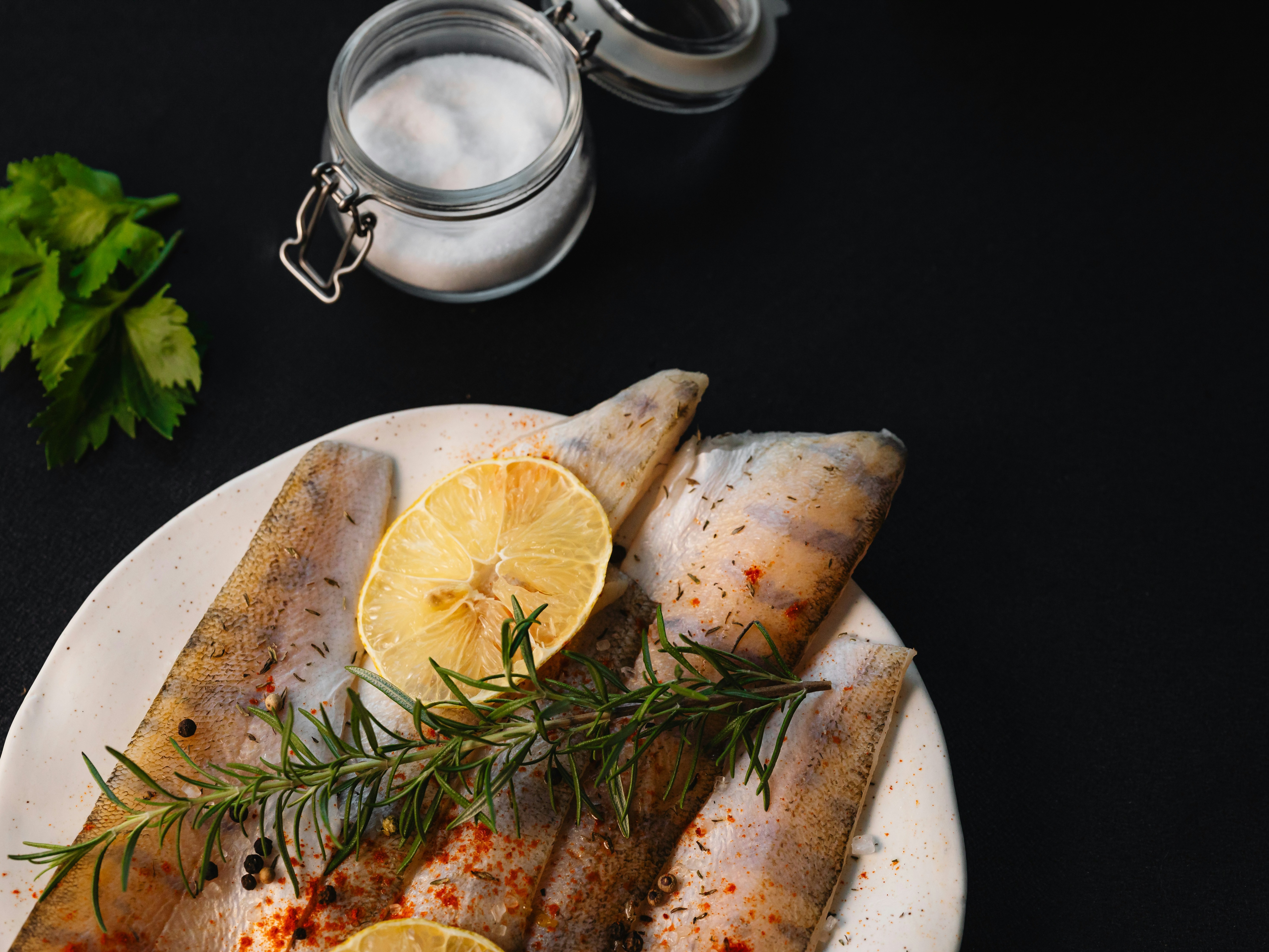The image shows three seasoned fish fillets on a white plate with a slice of lemon and sprigs of rosemary, accompanied by a jar of salt and parsley leaves on a dark background.