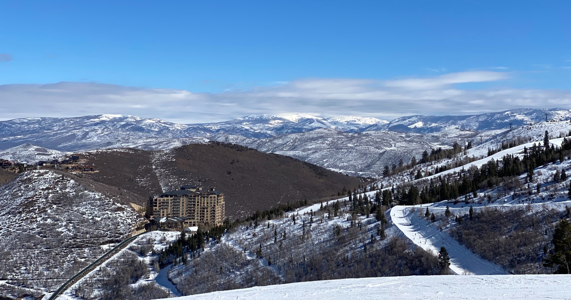 A view from the top of a snow covered mountain of Deer Valley and the resort at the bottom of the mountain on a partly cloudy day.
