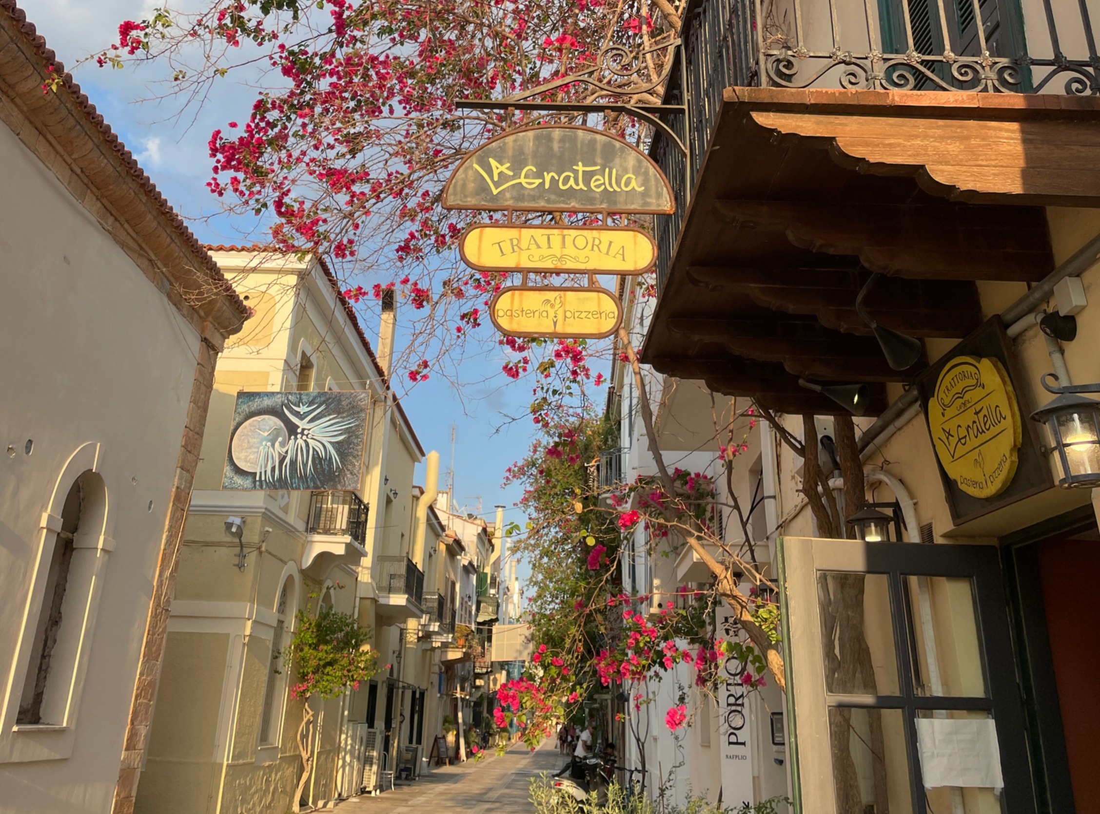 A quaint street with traditional buildings, a signed that reads "La Gratella Trattoria Pasteria Pizzaria" and a blooming tree under a clear sky.