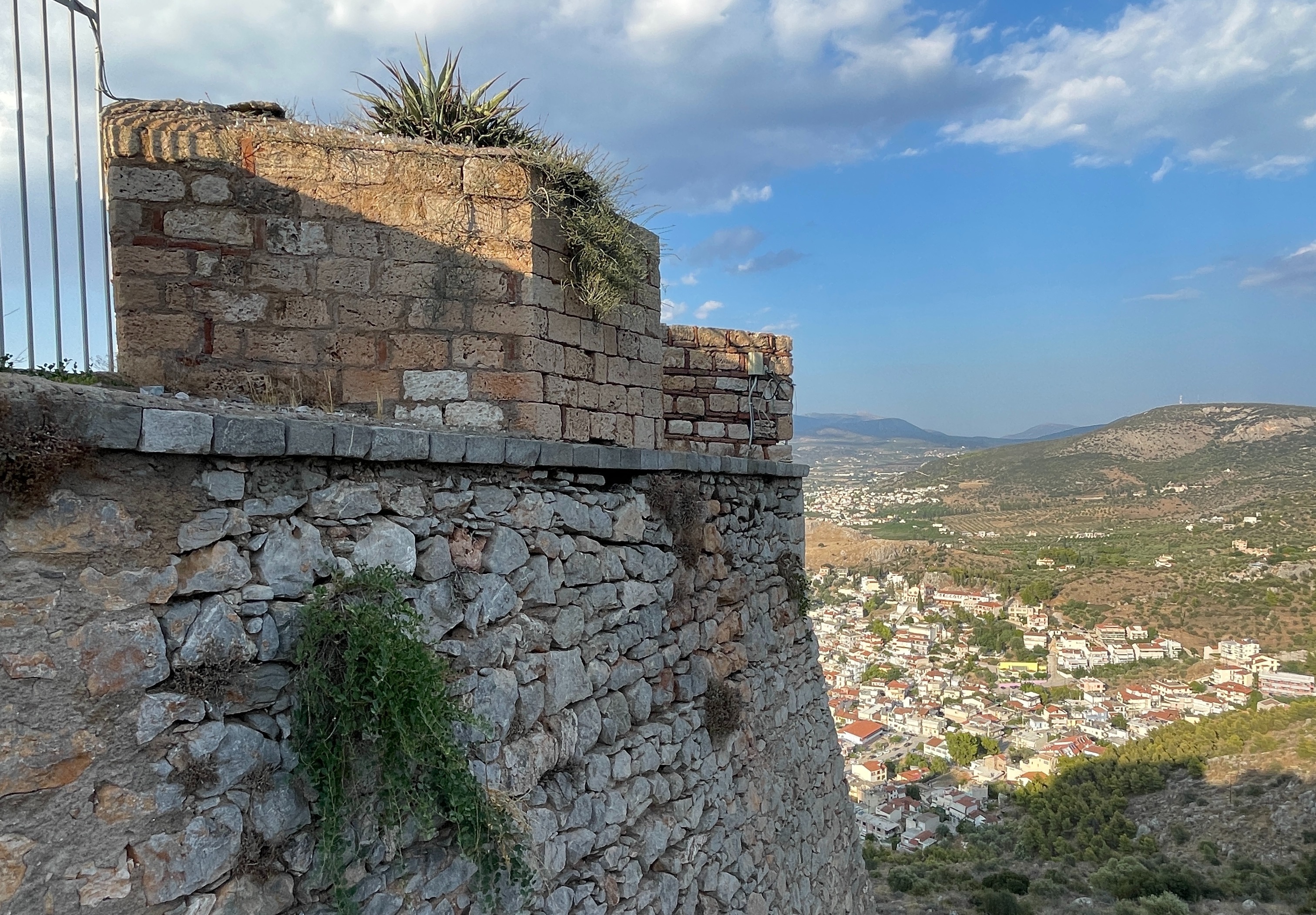 A stone structure with metal railings overlooks a scenic landscape under a blue sky with clouds.