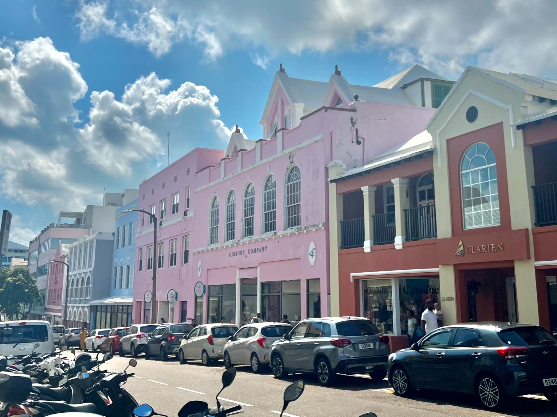A street view with colorful buildings, parked cars, and scooters under a blue sky with clouds.