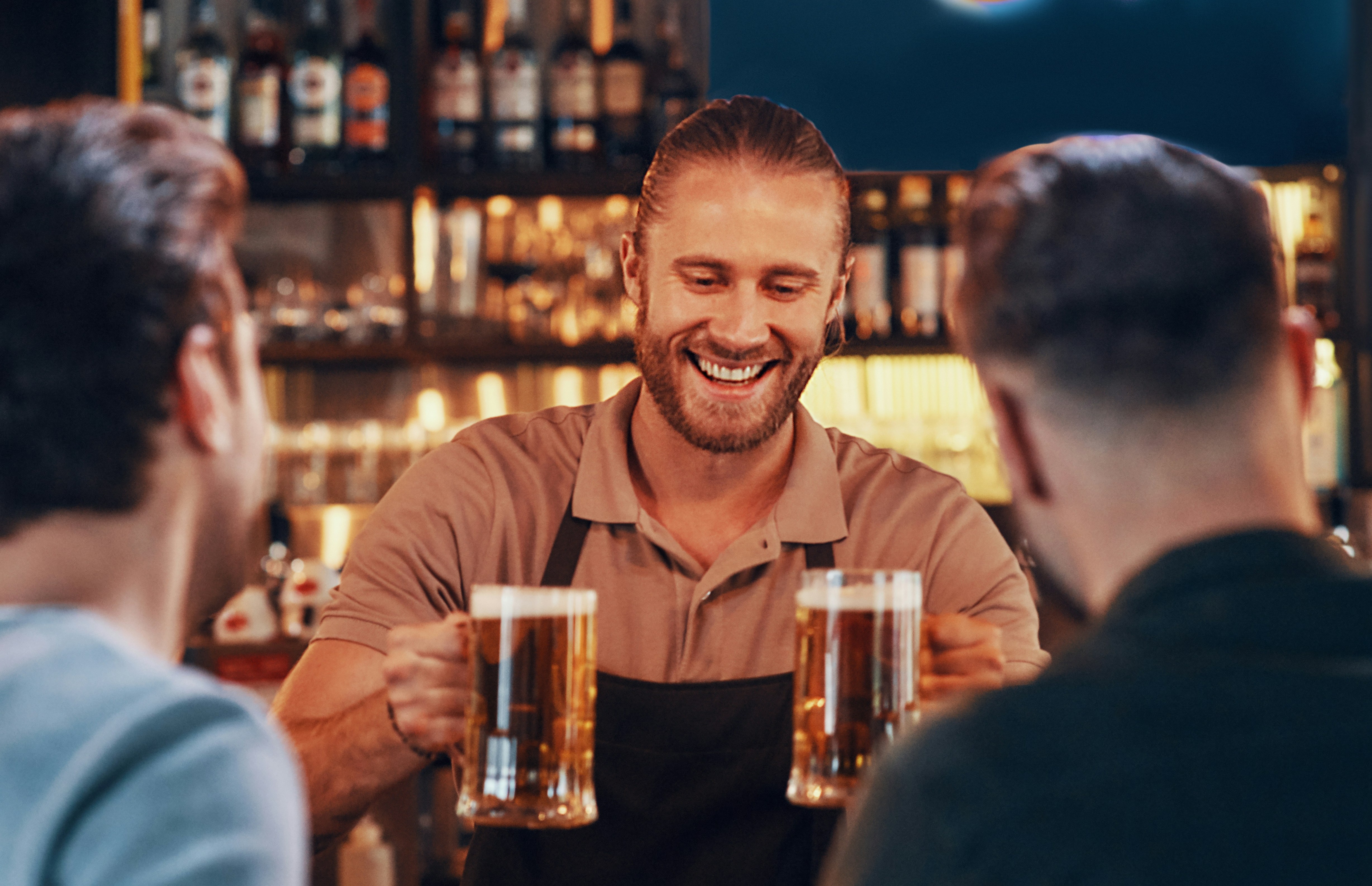 The image shows a bartender smiling, serving two pints of beer to customers at a bar.