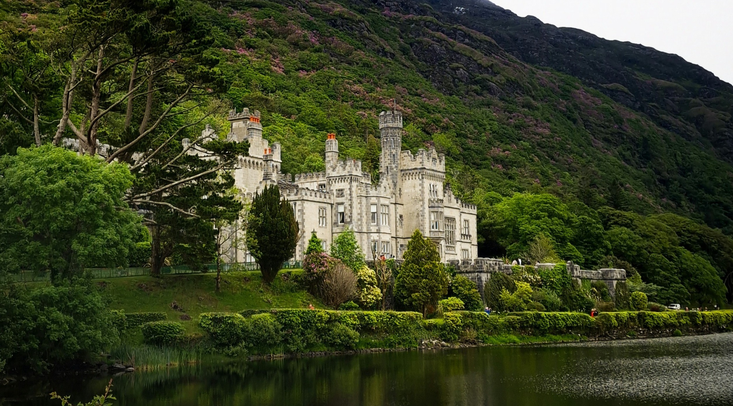 The image shows a grand castle with multiple turrets reflected in a calm body of water, surrounded by lush greenery and set against a backdrop of hills.