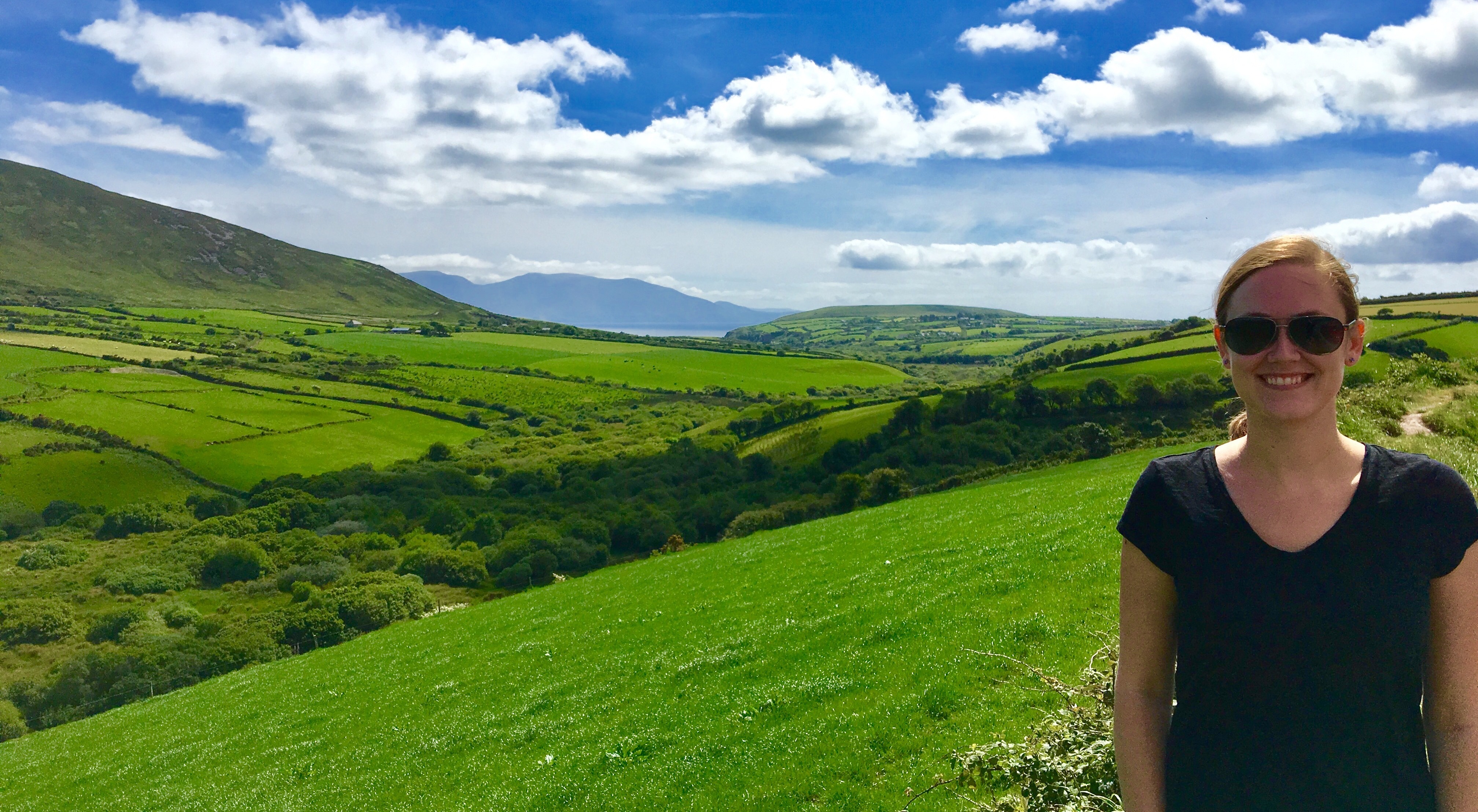 The image shows a person standing in a lush green field with rolling hills and a blue sky with clouds in the background.