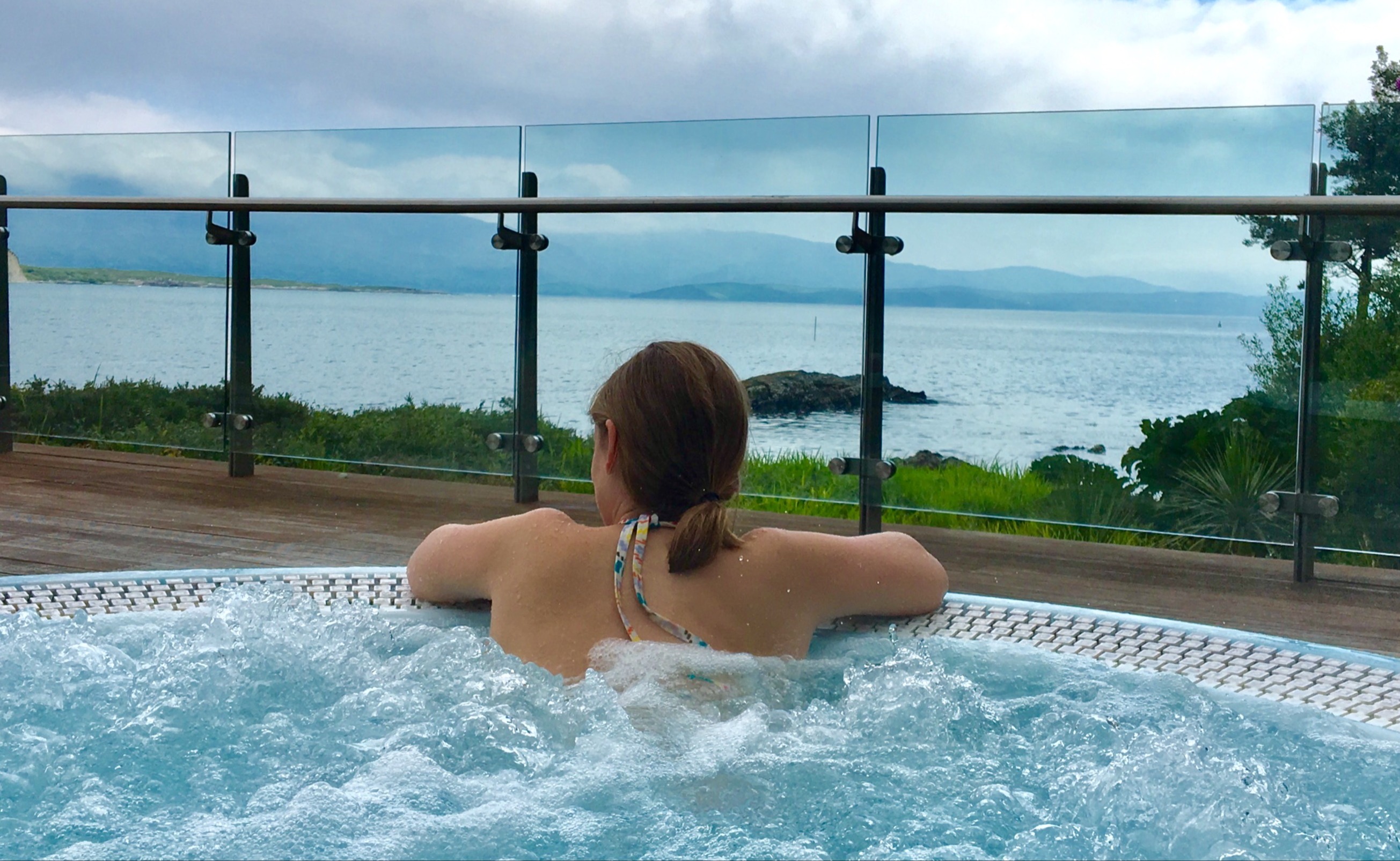 The image shows a person relaxing in an outdoor hot tub with a view of the sea and mountains in the background.