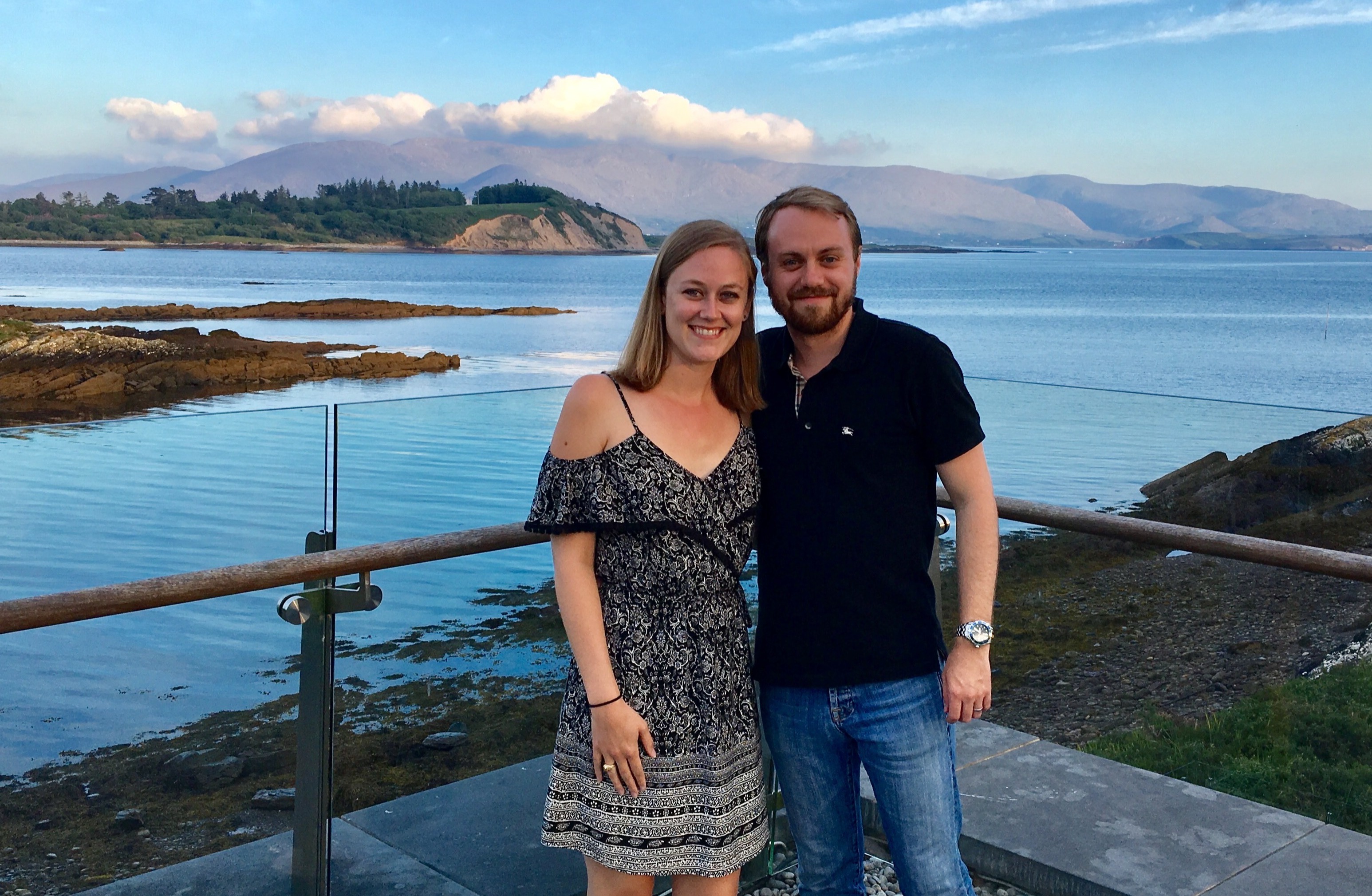 The image shows two individuals standing by a railing overlooking a scenic coastal landscape.