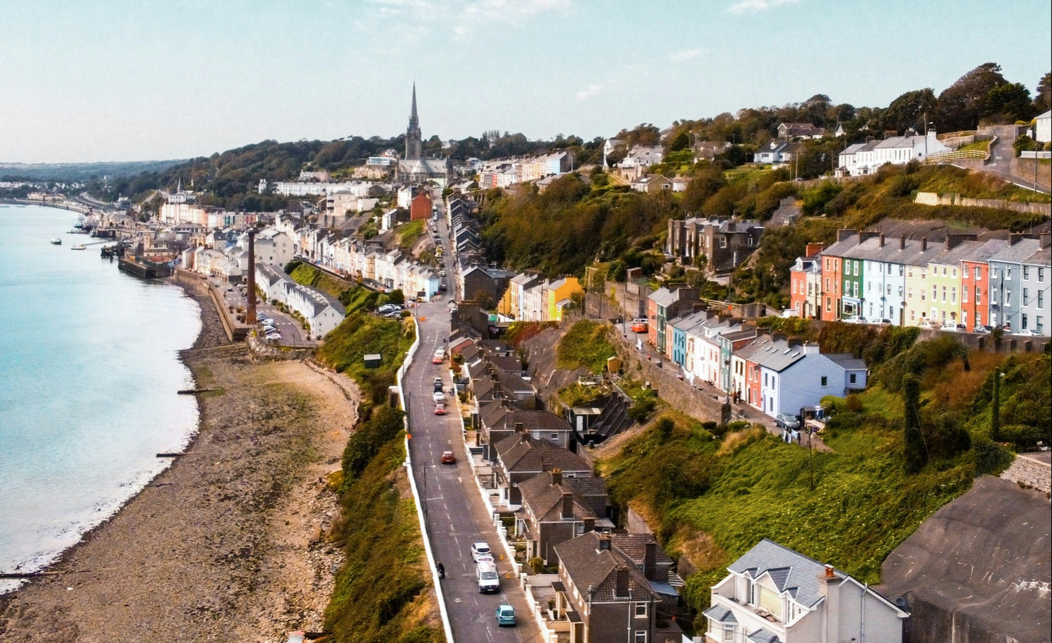 The image shows a coastal town with a clear blue sky, scattered clouds and a variety of buildings along the shoreline