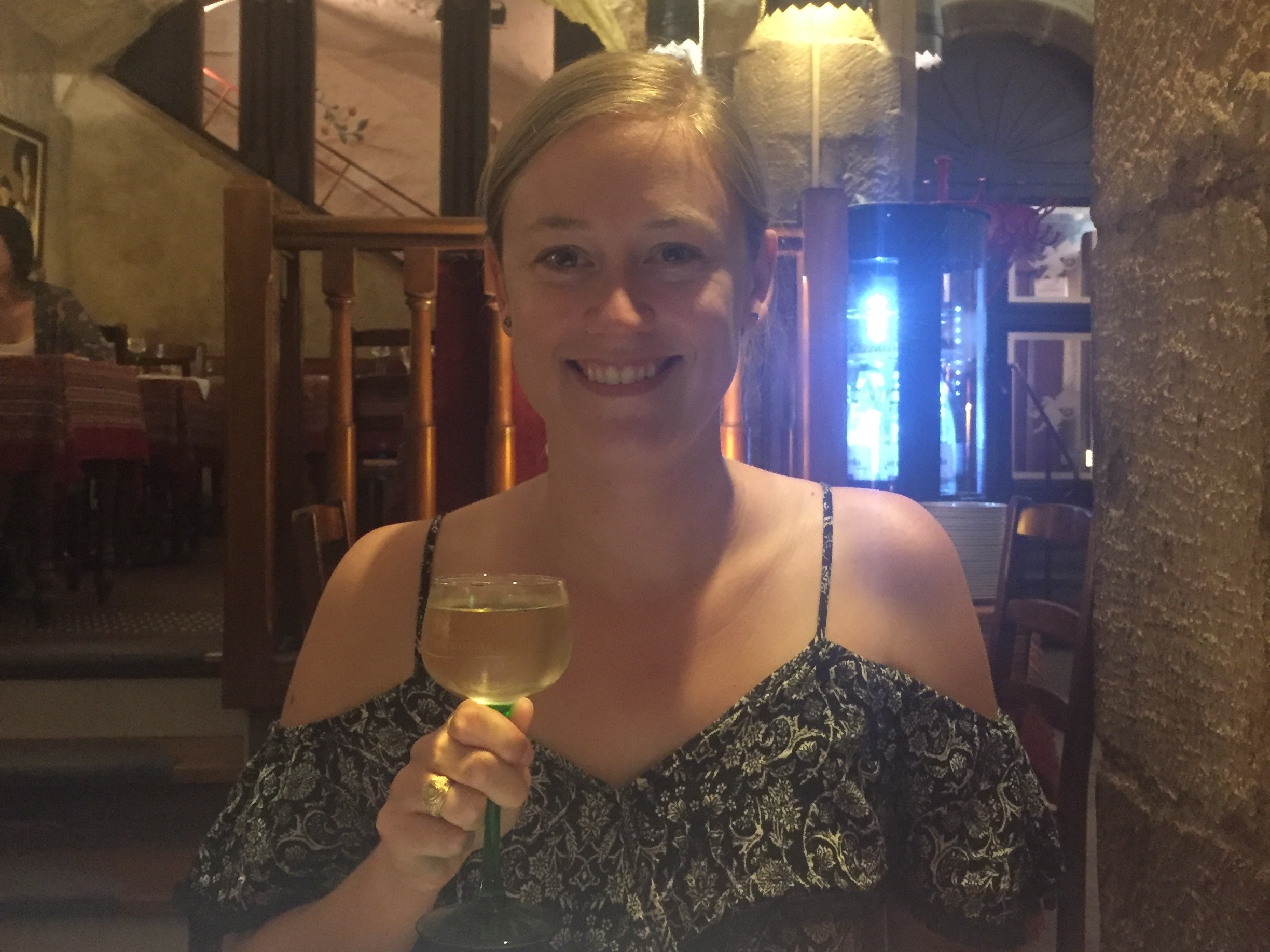The image shows an individual sitting at a table in a restaurant with arched ceilings, holding a glass of white wine.