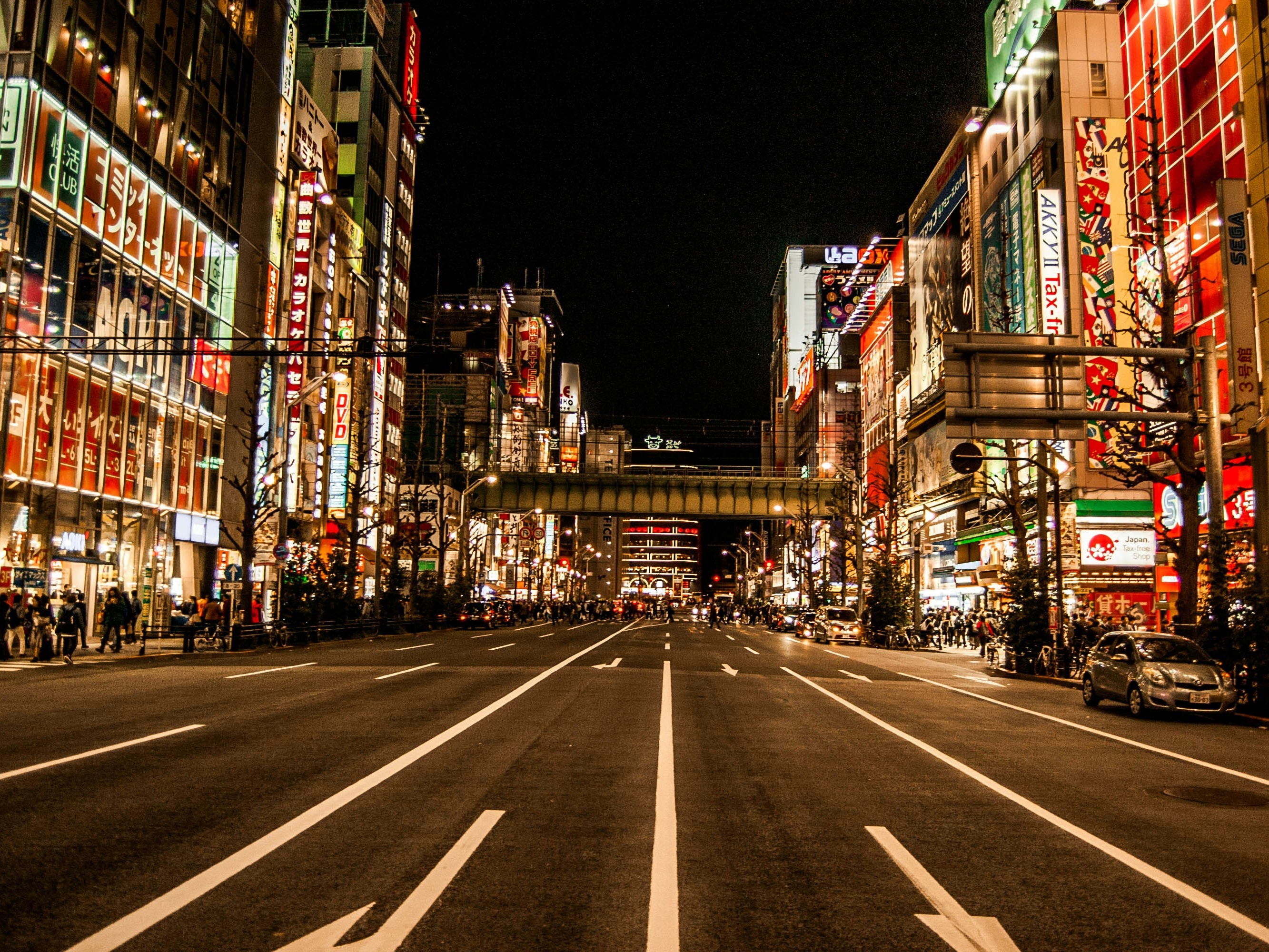 The image shows a bustling city street at night, illuminated by the lights of numerous signs and advertisements, with a pedestrian crossing in the foreground and an elevated train track in the distance.