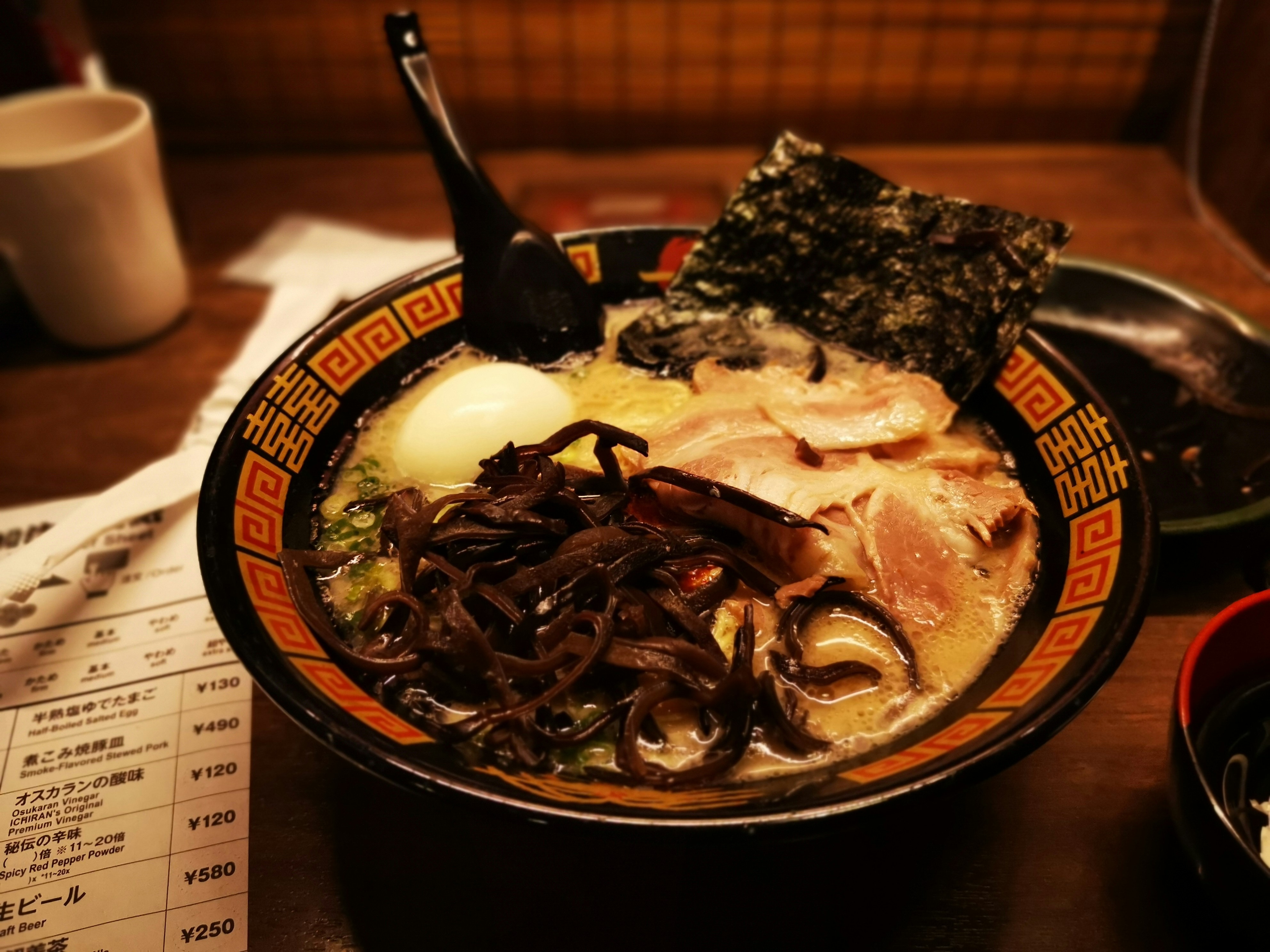 The image shows a close-up of a bowl of ramen with various toppings, set on a wooden table, suggesting a dining experience in a restaurant.