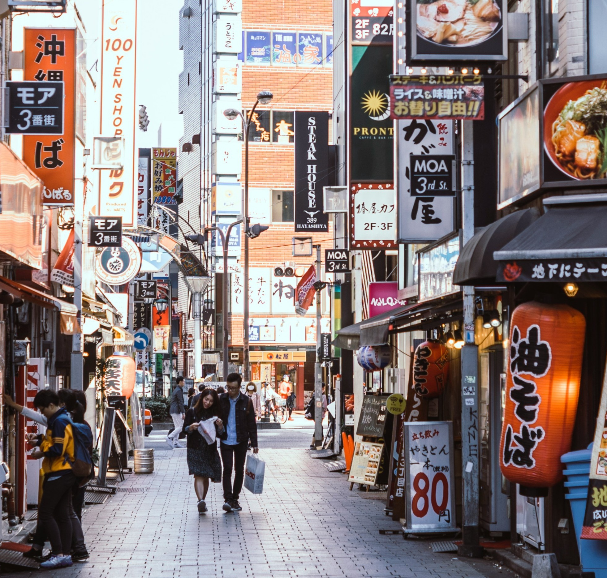 The image depicts a bustling street scene in Japan, lined with various shops and restaurants featuring colorful signage and advertisements.