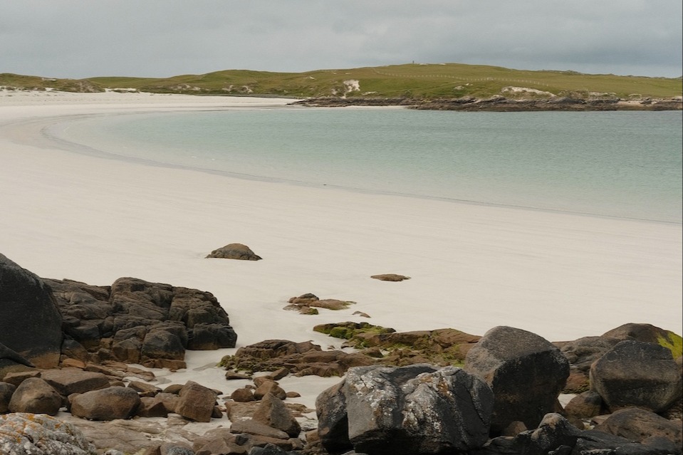 A calm ocean laps on the white sand shores of of Dog's Bay Beach on a grey day. Green hills in the distance.