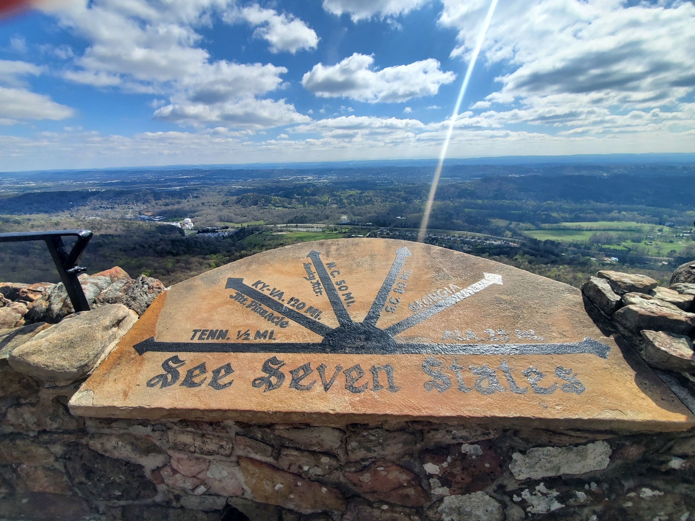 The image shows a scenic overlook with a stone compass indicating the direction and distance to various locations, under the phrase “See Seven States.”