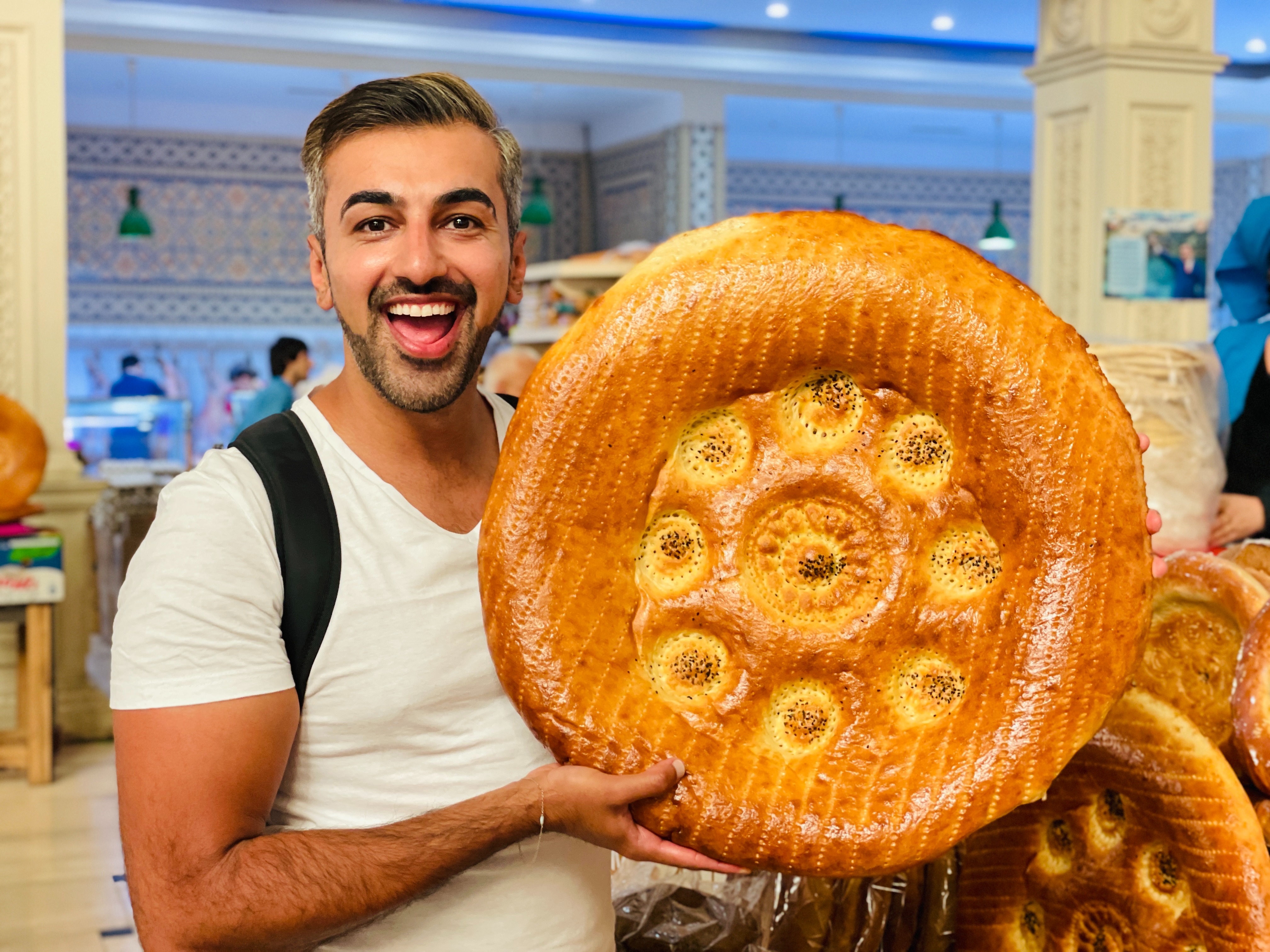 An individual holds a large, round bread with a patterned crust in a bakery or market setting.