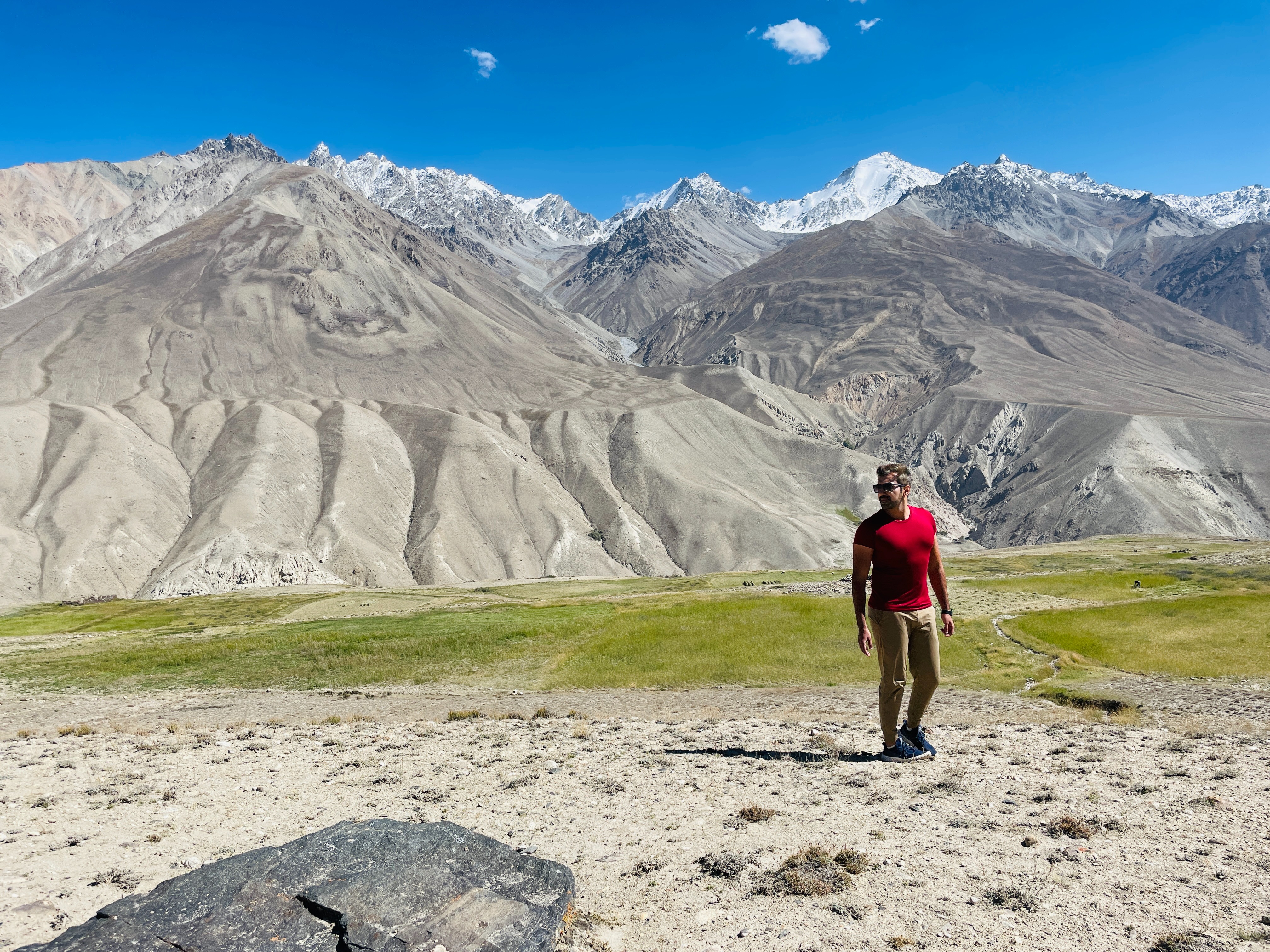 A person stands in a vast mountainous landscape, gazing towards the snow-capped peaks under a clear blue sky.