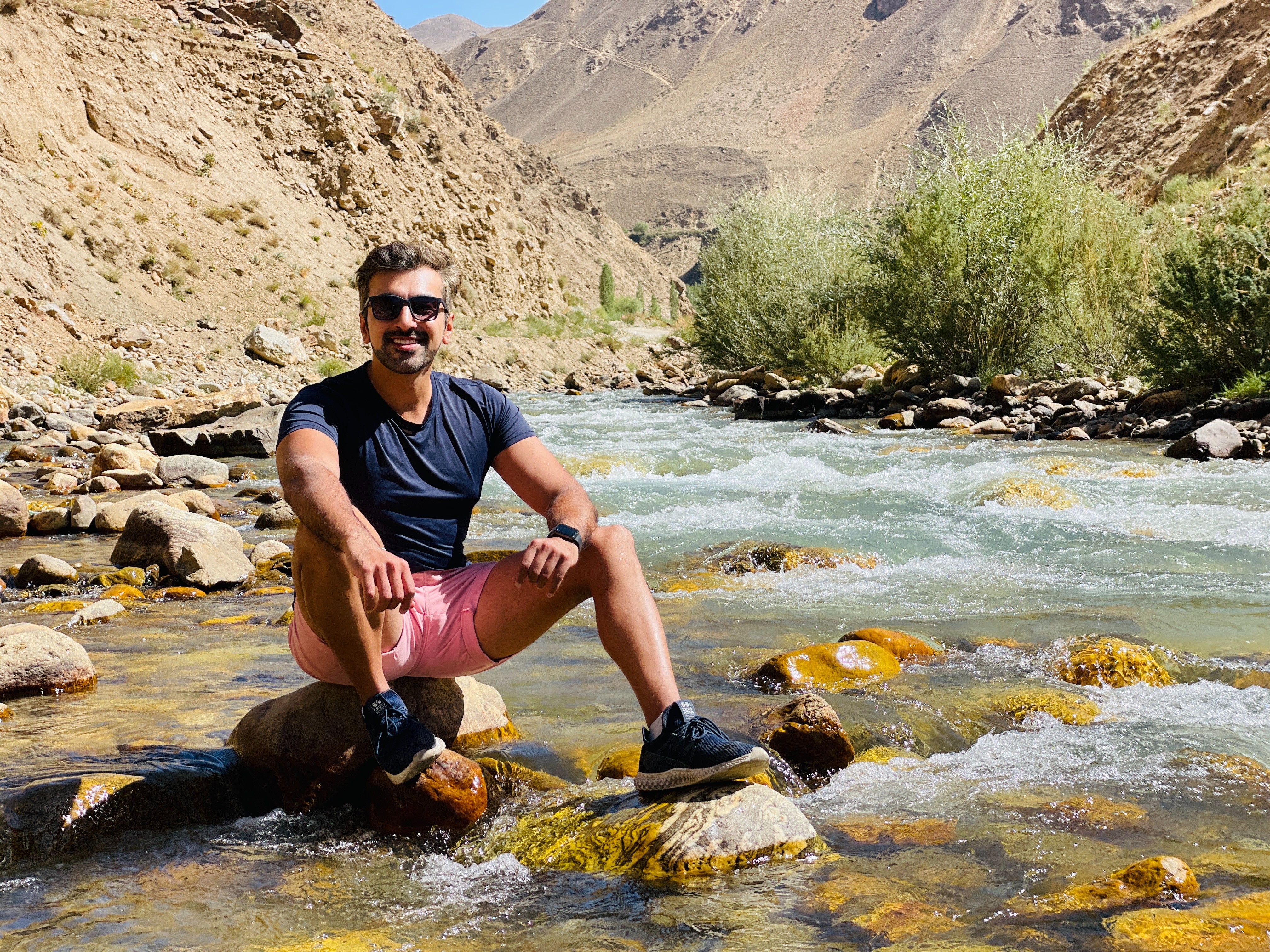An individual sits on a rock in the middle of a flowing river with a mountainous background.