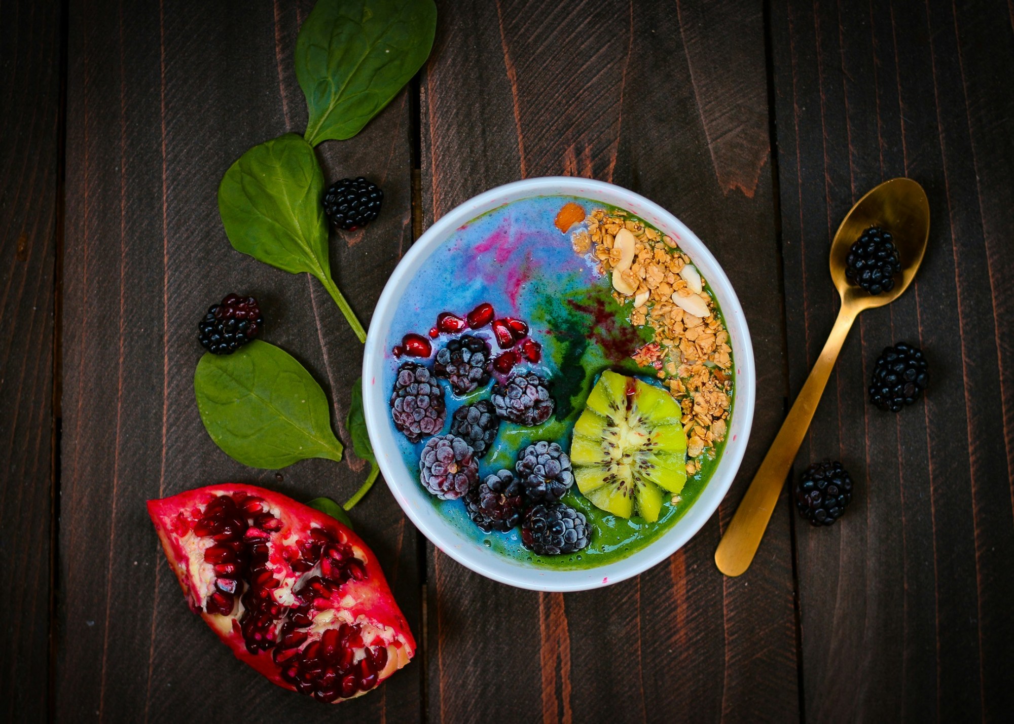 Granola, kiwi, blackberry and pomegranate on top of a blue smoothie in a bowl.