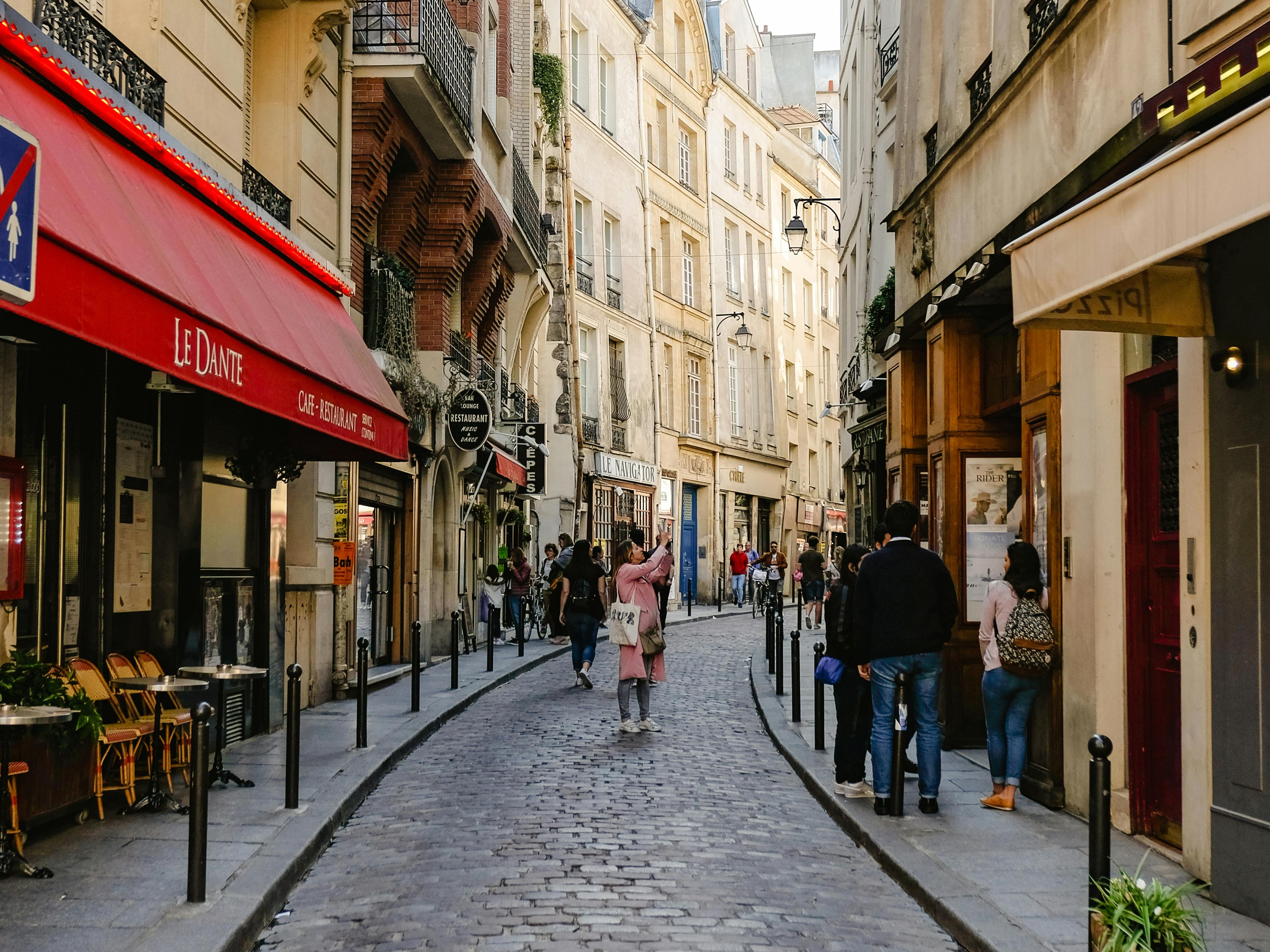 The image shows a narrow cobblestone street lined with buildings and small shops, bustling with pedestrians, capturing the charm of a European city.