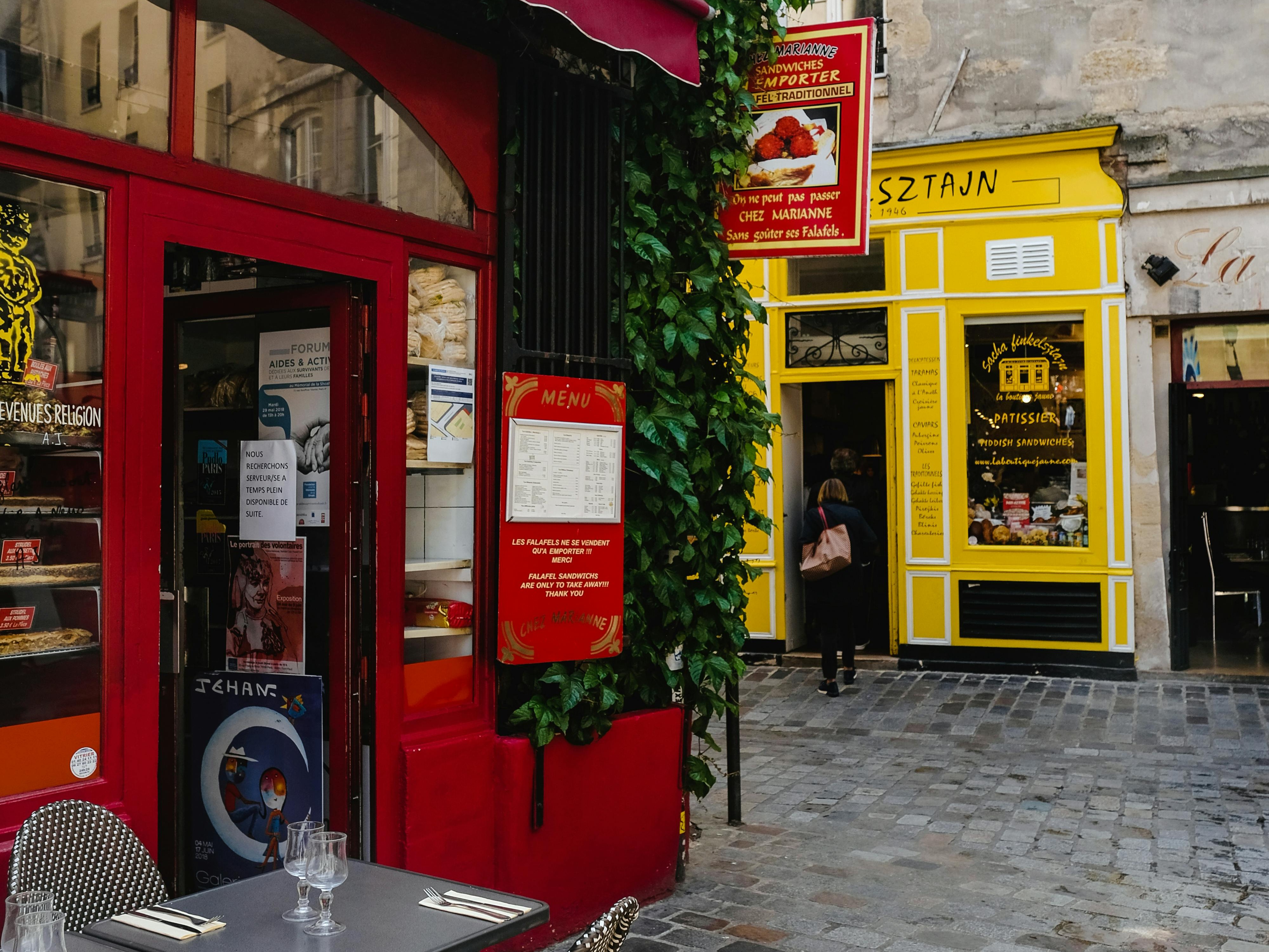 The image shows a vibrant street view with two contrasting storefronts, one red and one yellow, with outdoor seating and greenery.