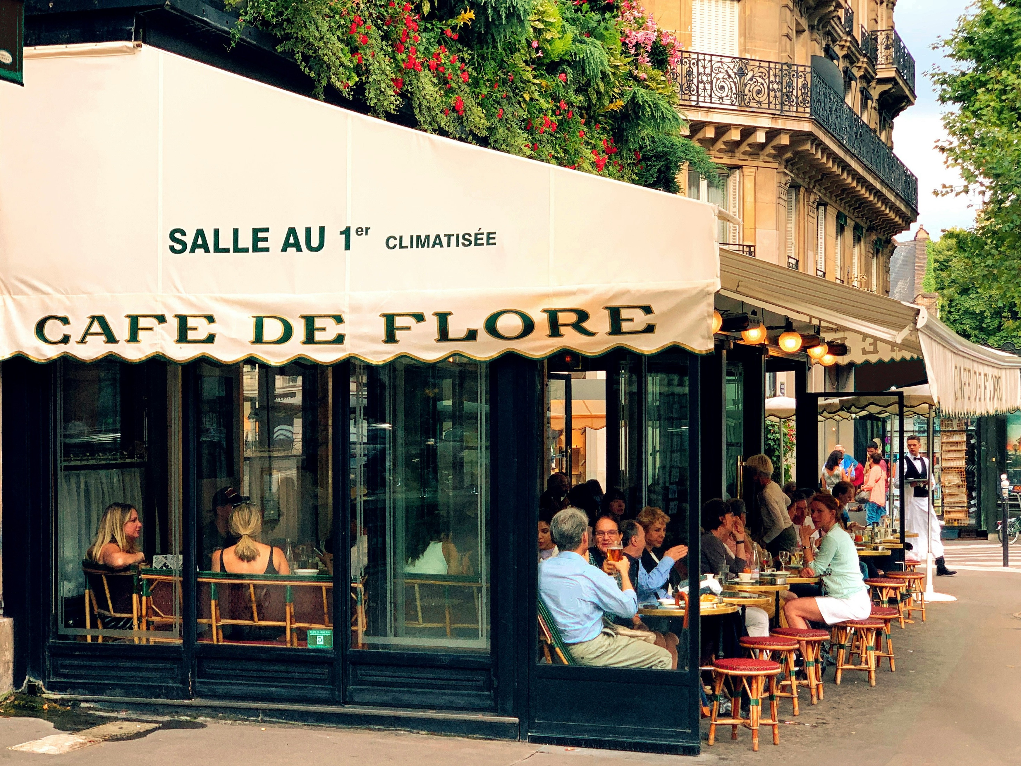 The image shows the exterior of Café de Flore, a bustling Parisian café with patrons seated outside and a sign indicating an air-conditioned room on the first floor.