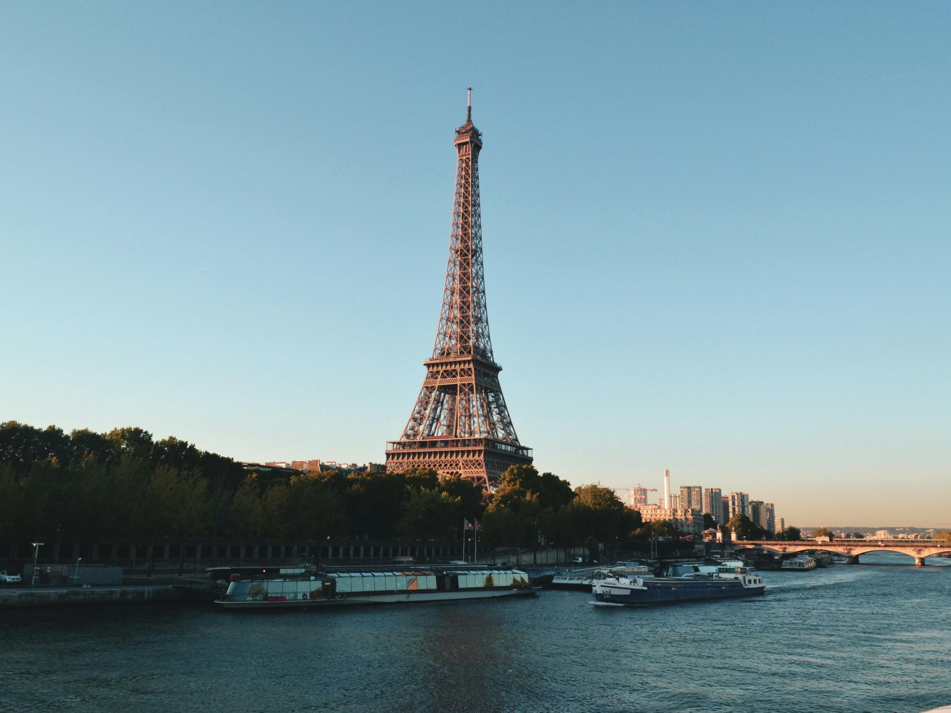 The image shows the Eiffel Tower beside the Seine River with a clear sky and a bridge in the background.