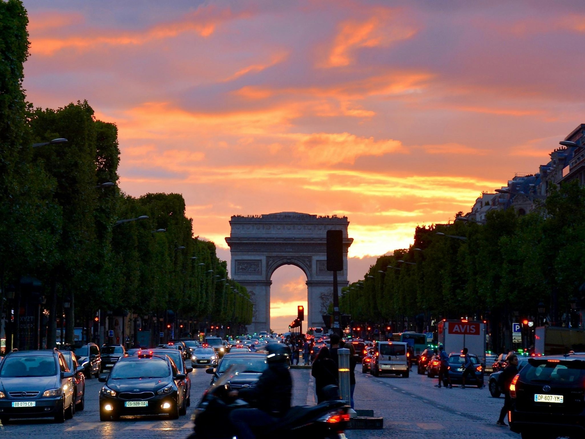 The image depicts the Champs-Élysées at twilight with the Arc de Triomphe in the background under a vibrant sunset sky.