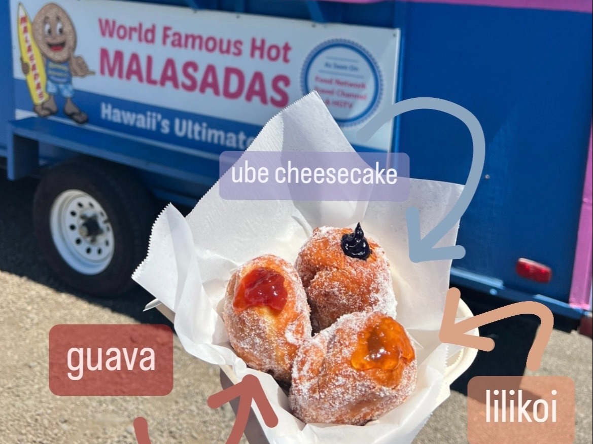 A hand holding three malasadas with different fillings in front of a colorful food truck.