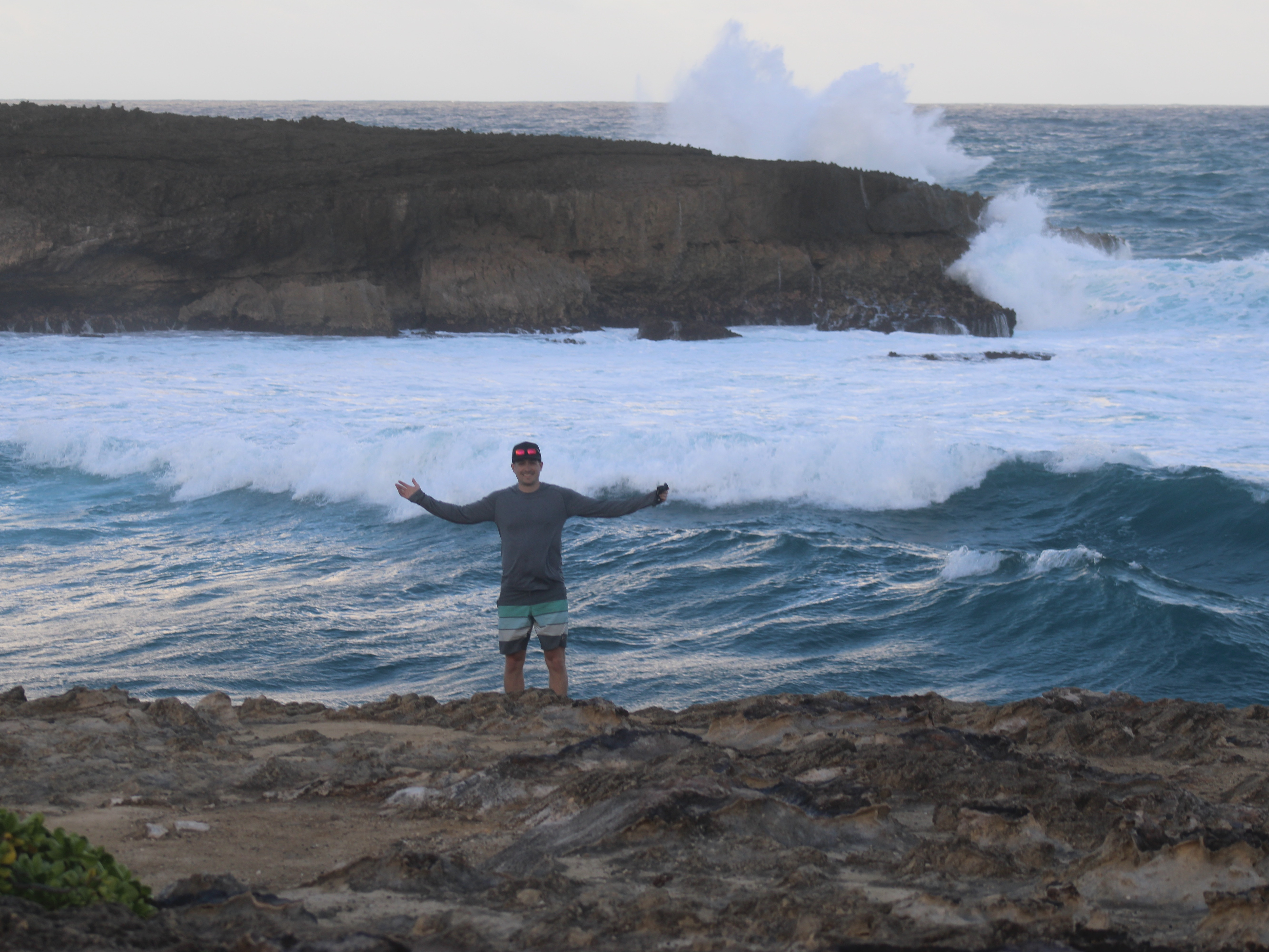 A person stands on rocky terrain with outstretched arms facing large waves crashing against the shore.