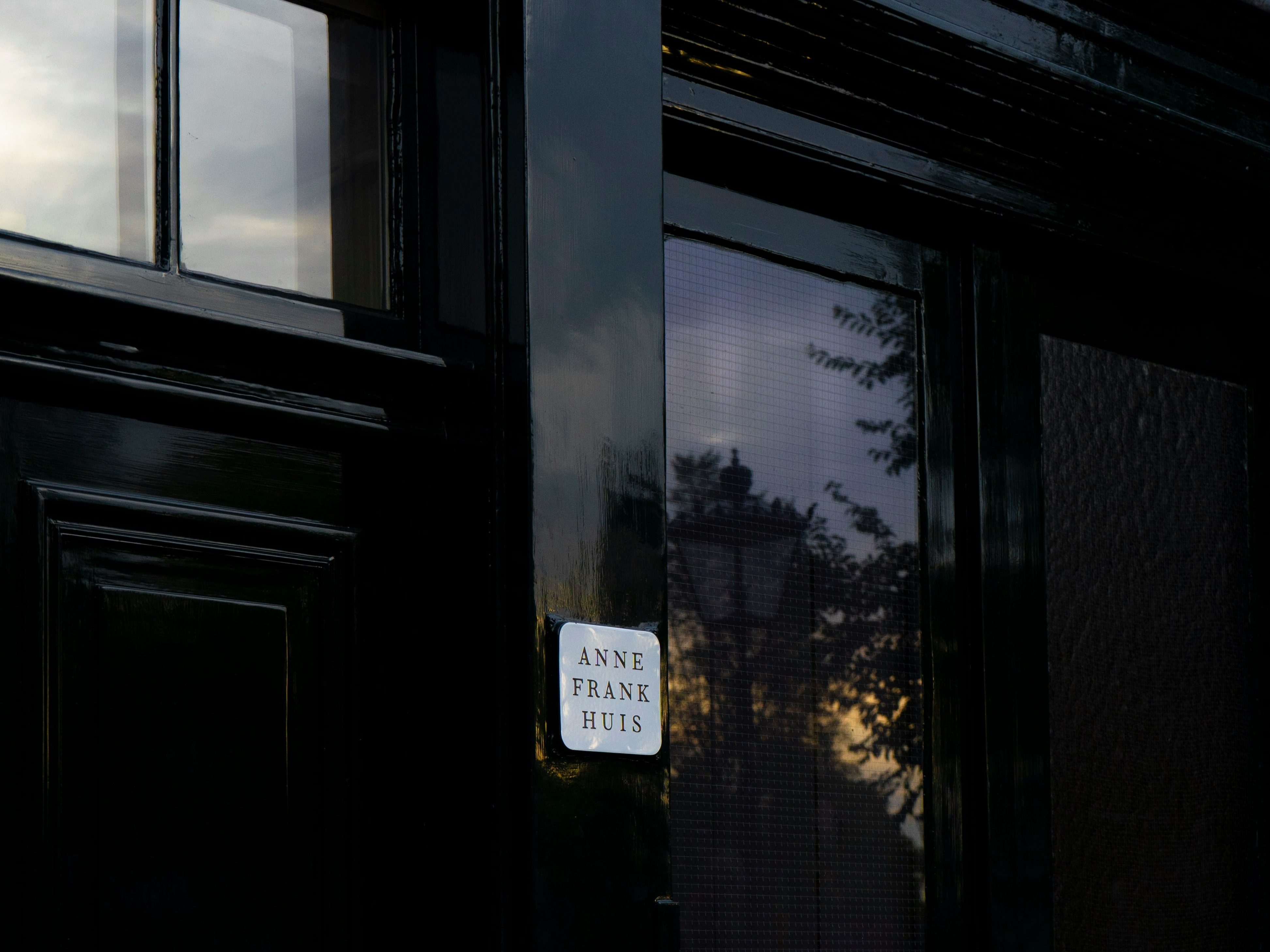 A dark-colored door with a sign that reads “Anne Frank Huis,” reflecting the silhouette of trees and a sunset.