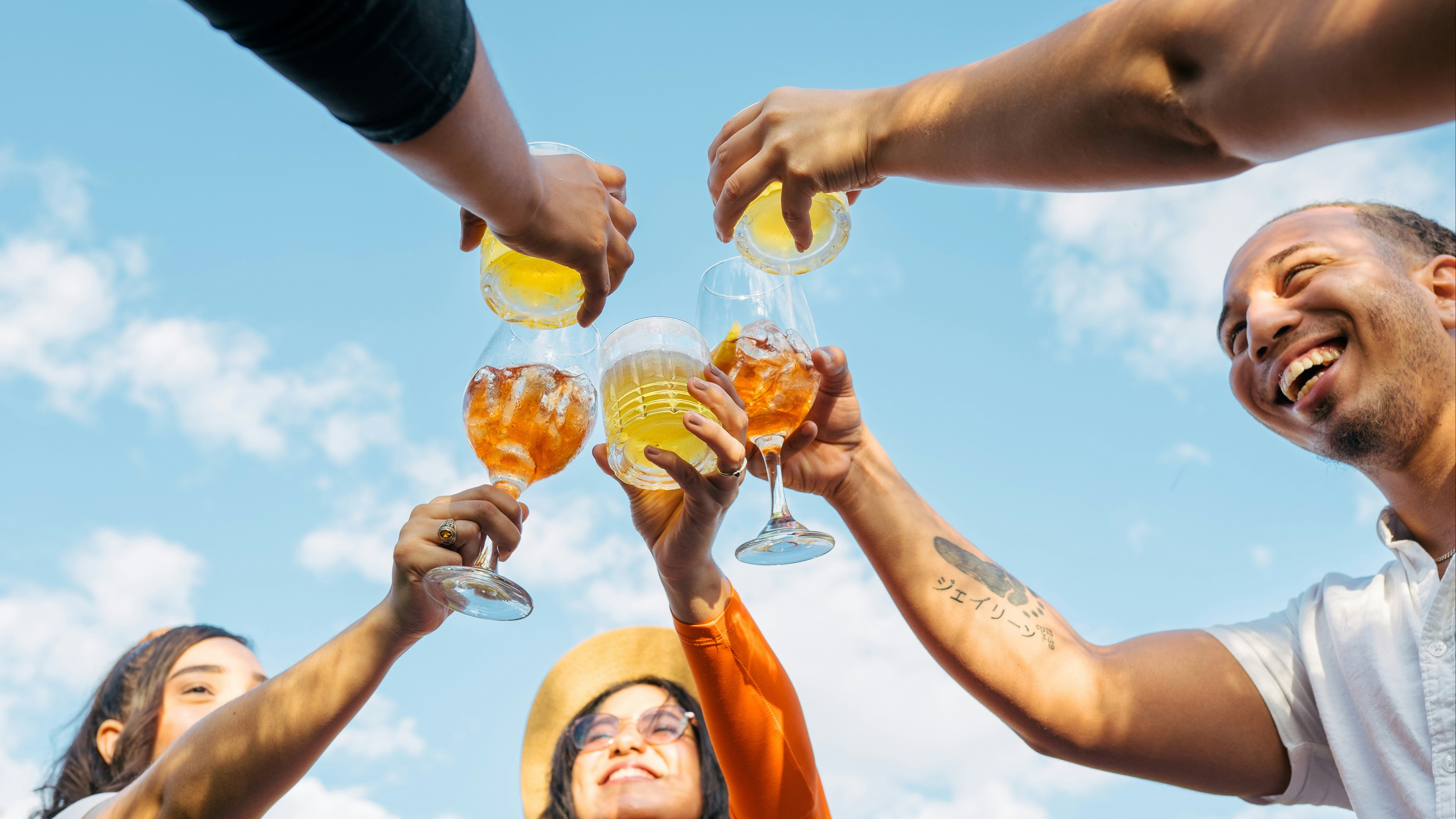 A group of individuals are toasting with drinks against a blue sky, with their faces obscured by brown rectangles.
