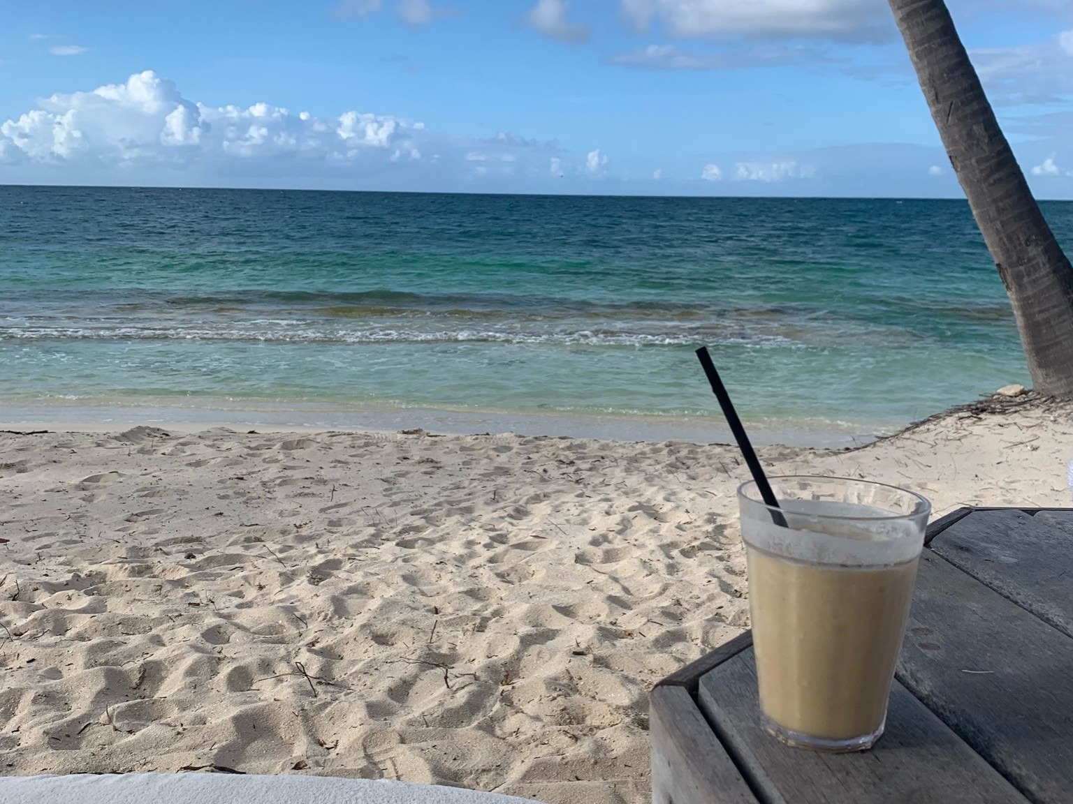 A tranquil beach scene with a clear sky, calm sea, sandy shore, a beverage on a table and part of a sunbed under the shade.
