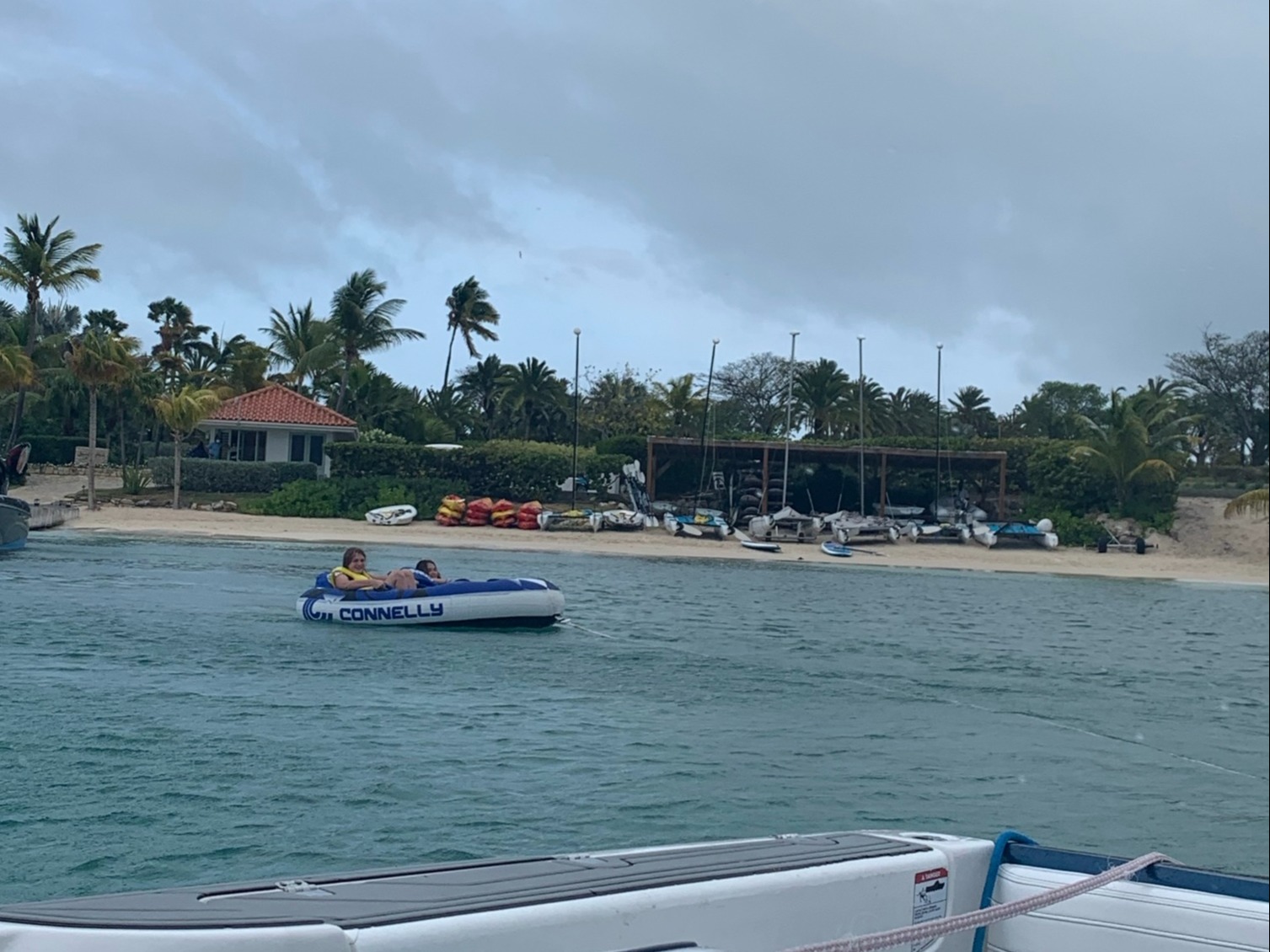 A view from a boat showing another boat and shoreline with palm trees and buildings under an overcast sky.